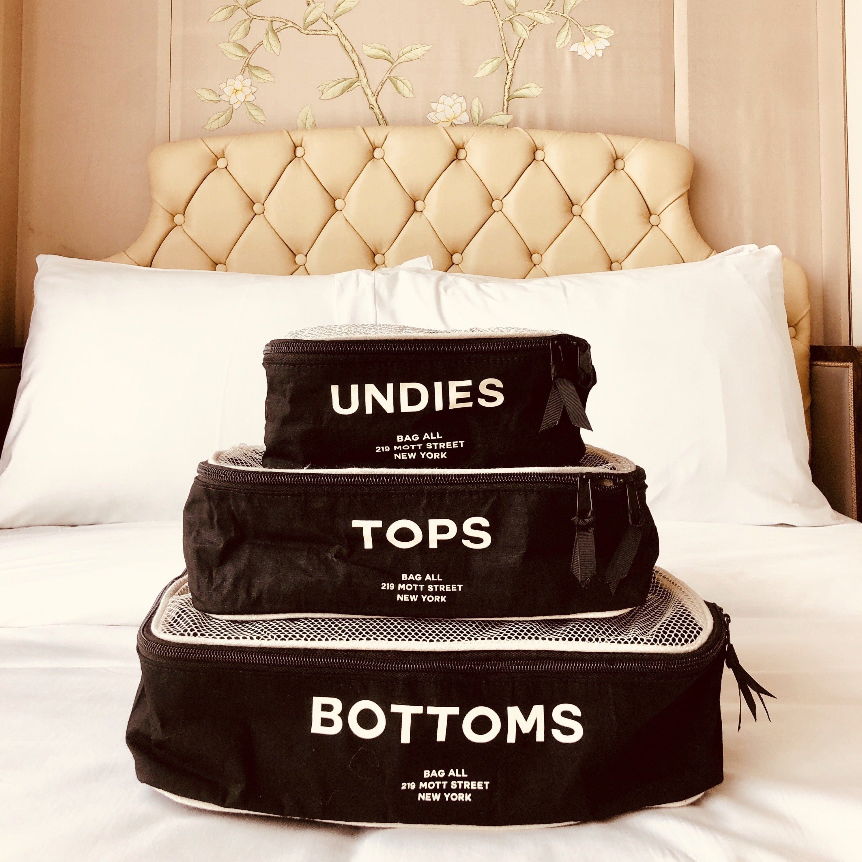 Bag-all Cotton Packing Cubes 3-pack in Black with white mesh and text labels for Undies, Tops, and Bottoms, stacked on white bed against tufted headboard