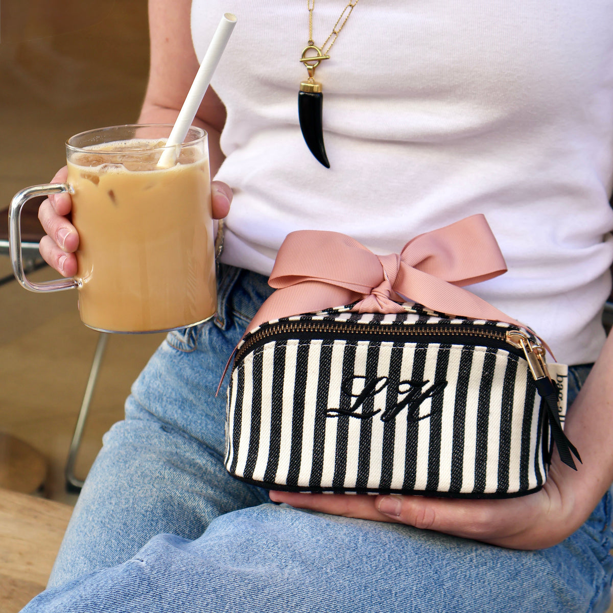 Bag-all striped travel pouch with a pink bow held on a travelerโs lap beside iced coffee