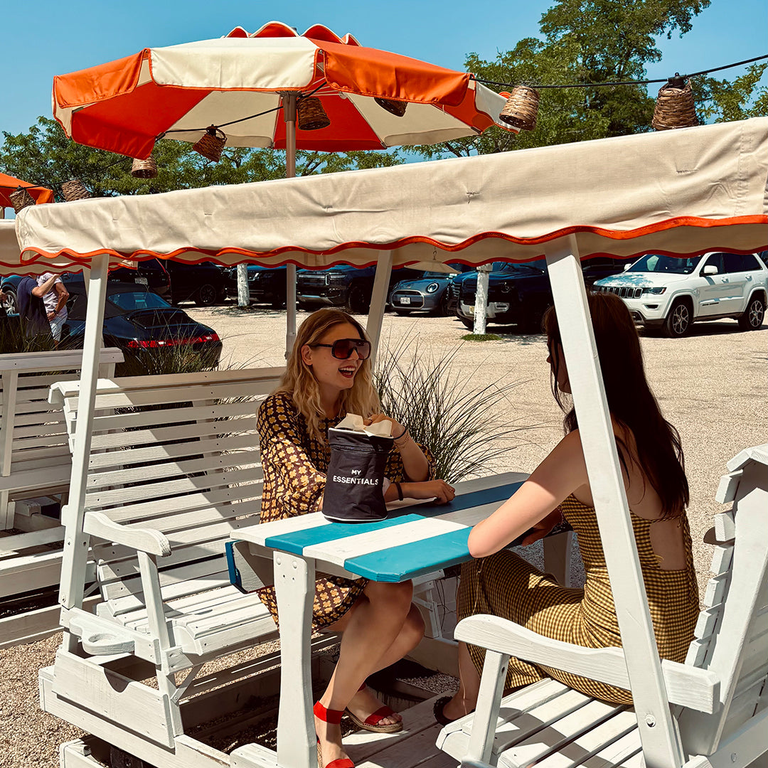 Two women sit under an orange and white canopy enjoying a sunny day with a black Bag-all essentials pouch on the table