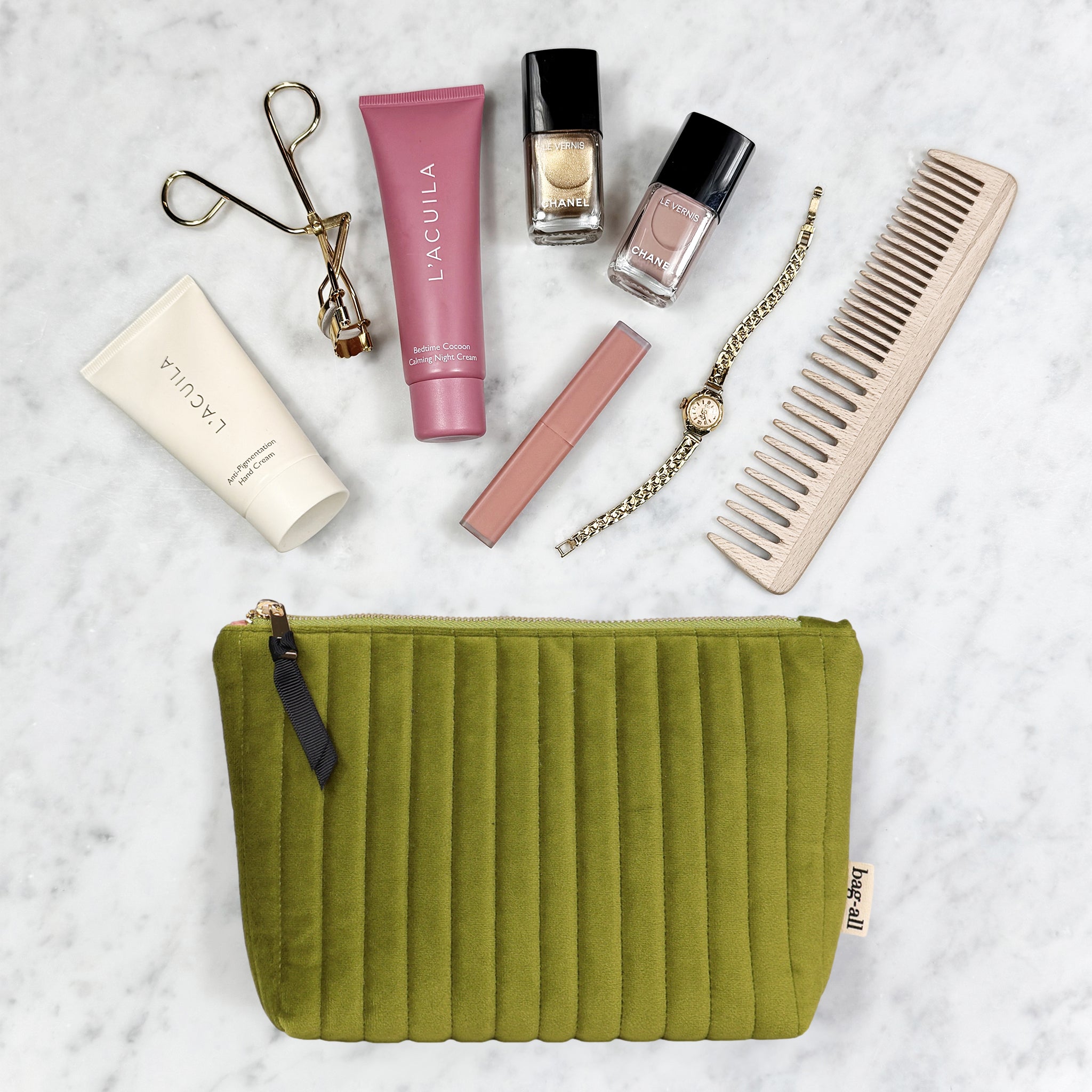 Quilted green velvet Bag-all makeup pouch with cosmetics, nail polish, comb, and watch on marble surface
