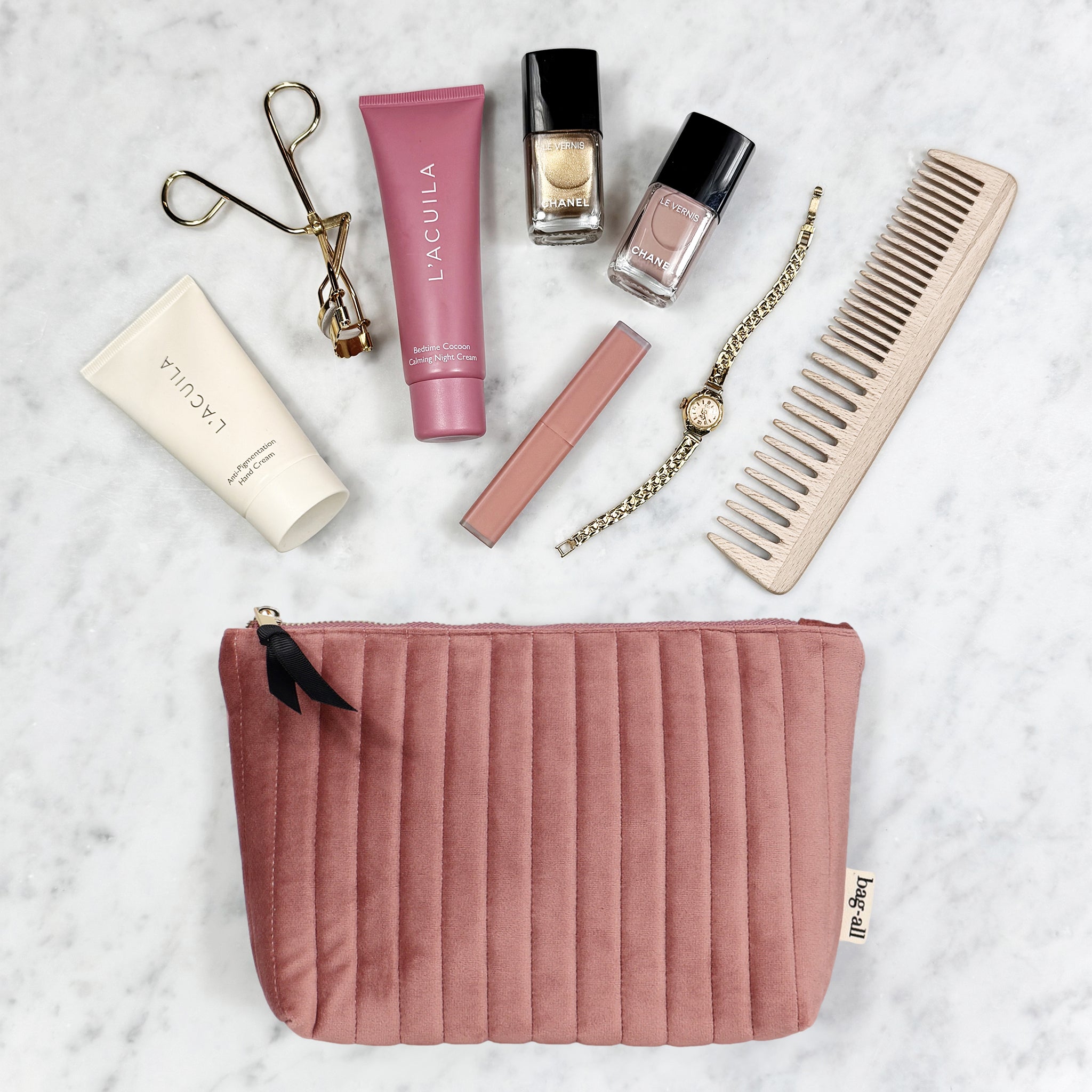 Pink quilted velvet Bag-all makeup pouch with beauty essentials and gold watch on marble surface