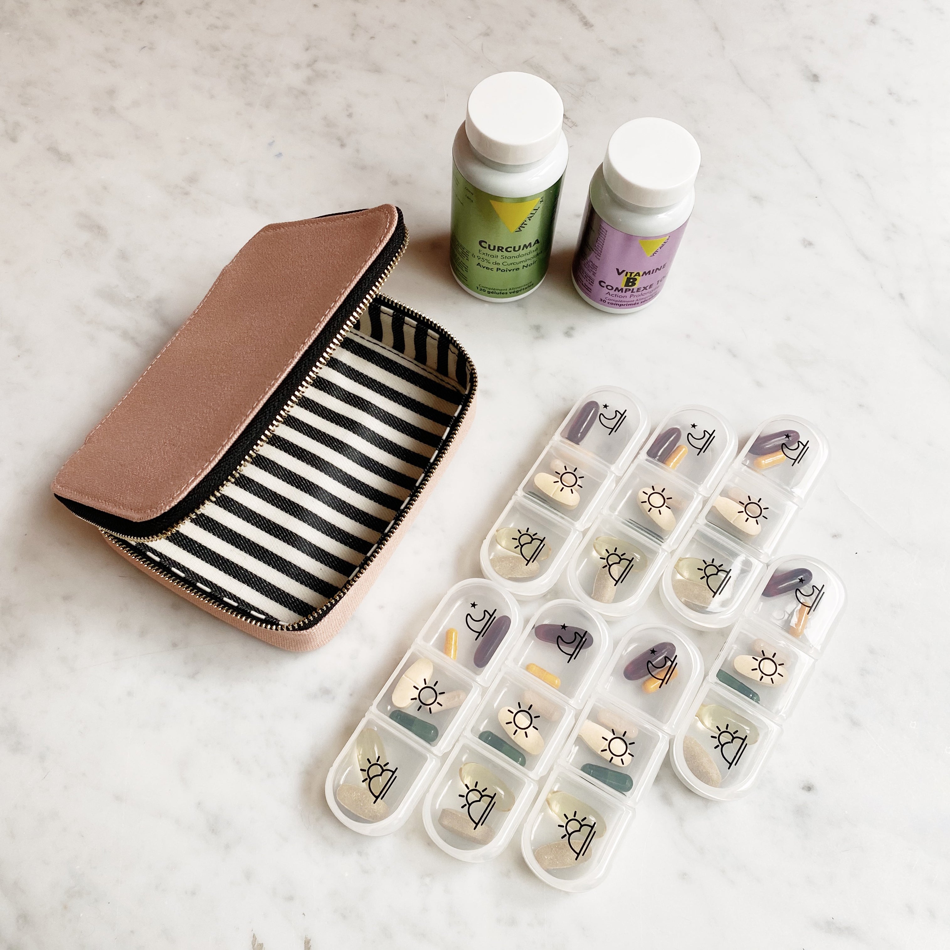 Bag-all pink blush vitamin organizer with striped interior, daily pill compartments, and supplement bottles on marble surface