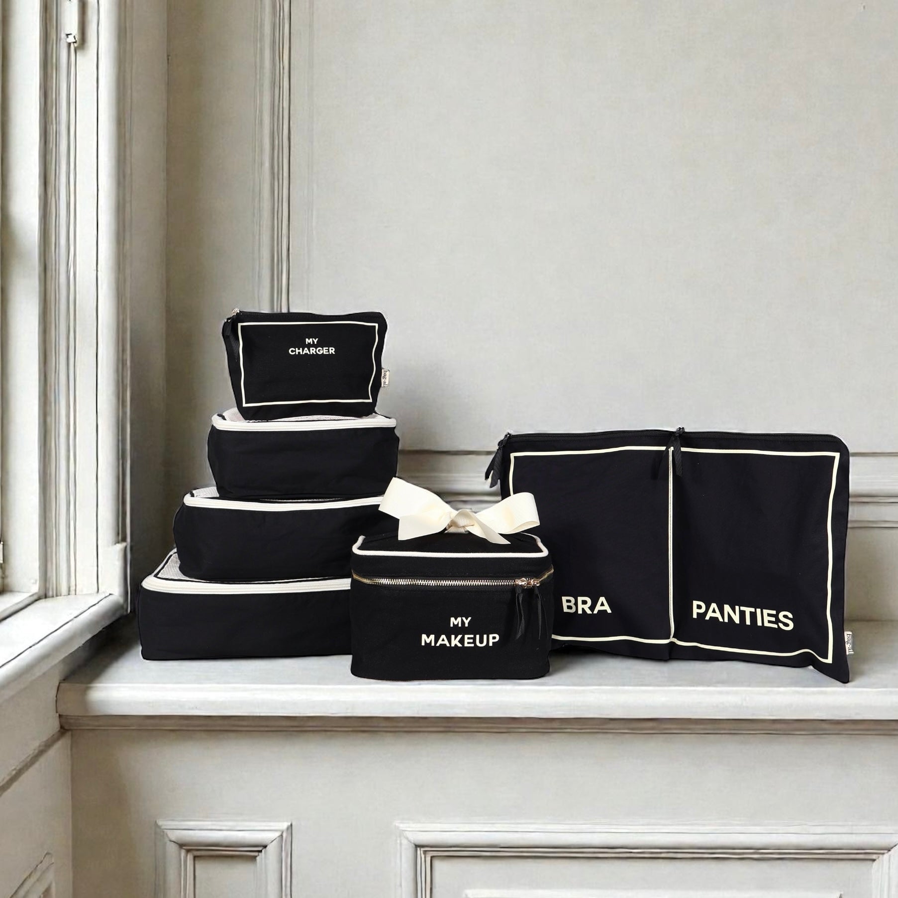 Black Bag-all Travel Glam 4-Pack with labeled makeup, charger, bra, and panties organizers on white ledge near window
