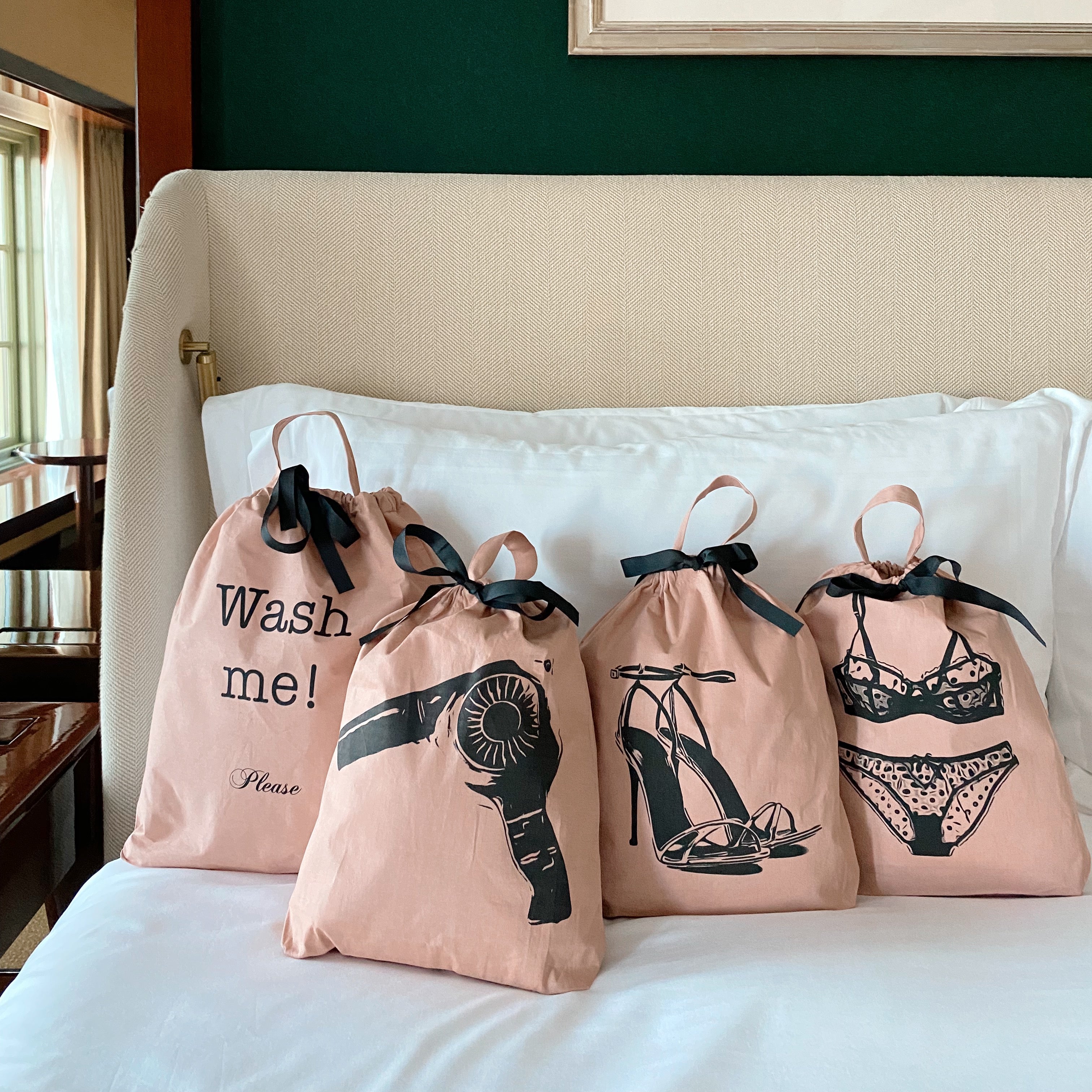 Bag-all pink travel organizer bags with black drawstring and prints featuring hairdryer, wash me, shoes and lingerie designs on cream couch, perfect for travel storage and organization