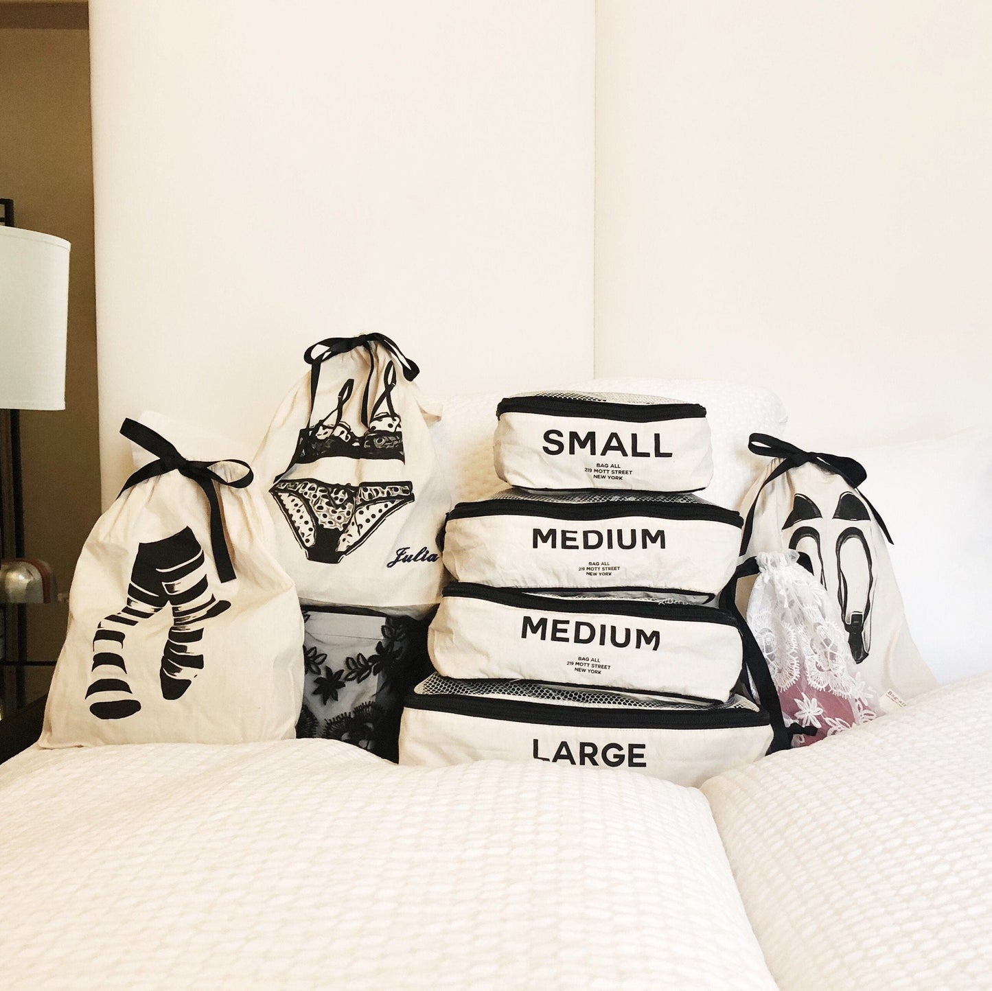 Bag-all Sock Short Bag in Cream - Organized storage solution featuring stackable cotton bags with size labels and black ribbon ties, displayed on white bedding alongside lingerie and sock bags