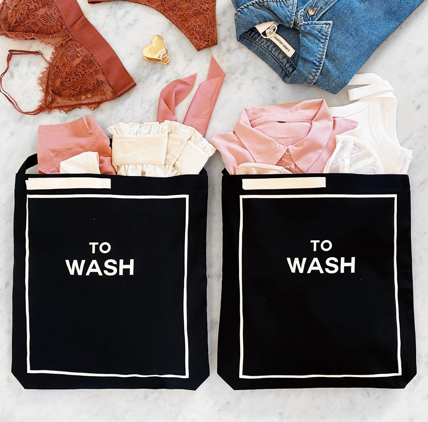 Bag-all To Wash Laundry Bag in black cotton with white border detail and text, shown filled with pink and neutral clothing items on marble surface, perfect for organizing dirty laundry