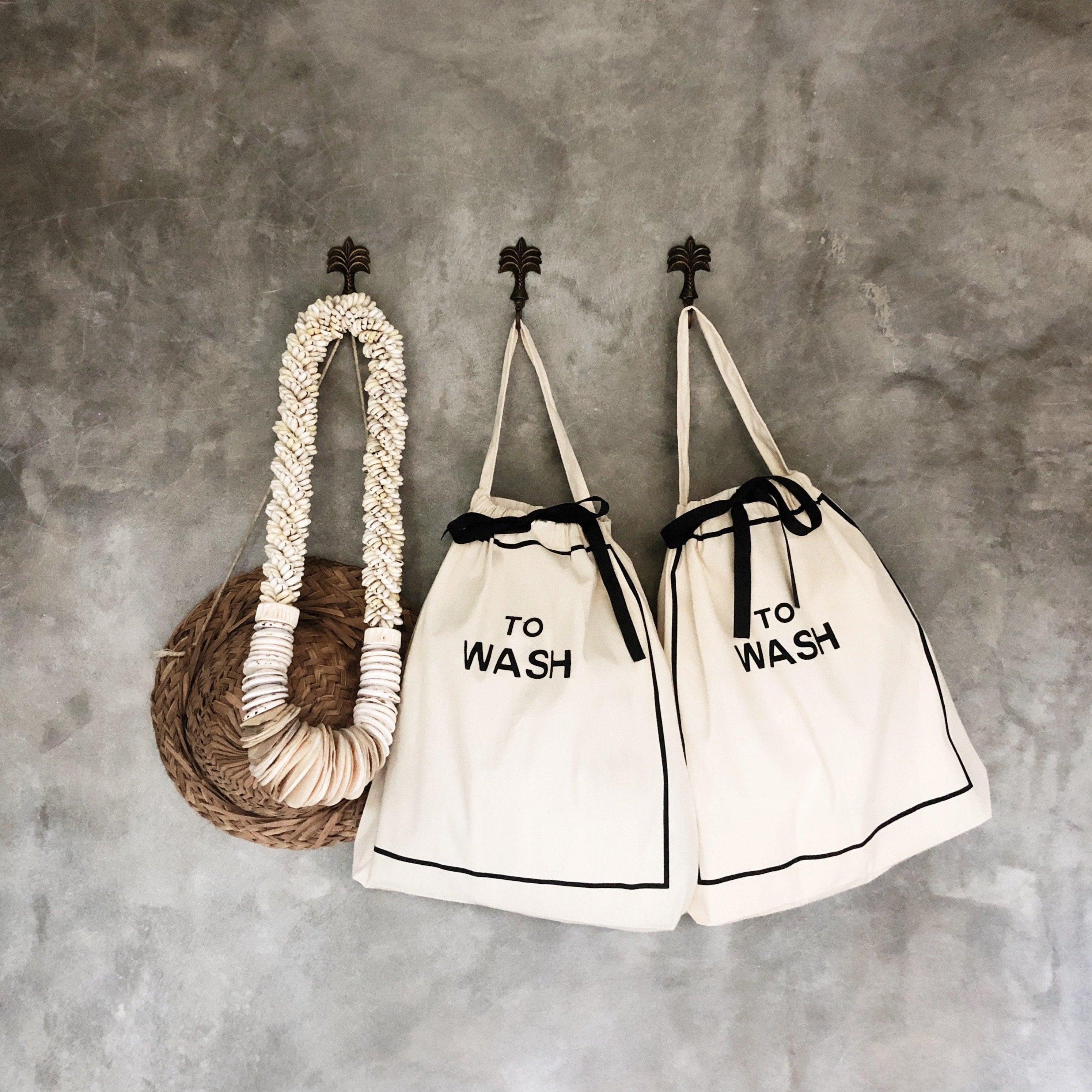 Bag-all To Wash Laundry Bag in cream cotton with black text, featuring drawstring closure and hanging handle, displayed alongside matching bag against textured gray wall