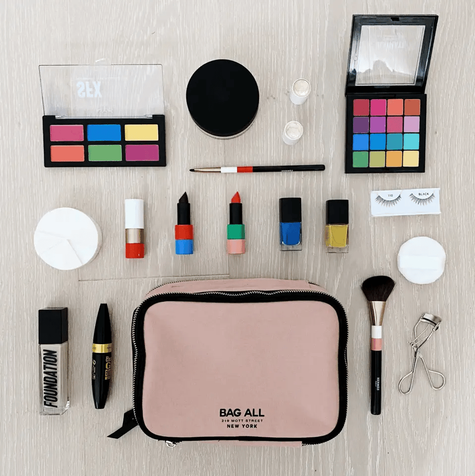 Bag-all Double Sided Toiletry Case in Pink/Blush surrounded by makeup essentials including eyeshadow palettes, lipsticks, nail polishes, brushes, and beauty tools on light wooden surface