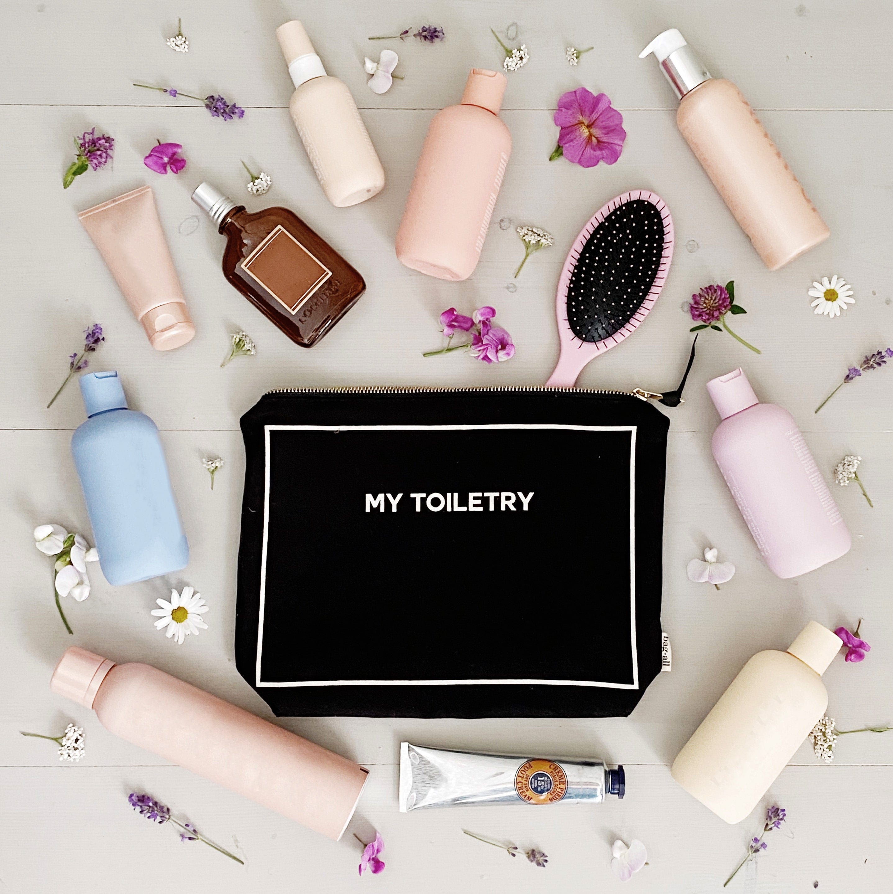 Bag-all Weekend Essential Packing Set: Black cotton toiletry pouch surrounded by pastel beauty products, hairbrush, and delicate flowers on light wooden surface, showcasing travel organization