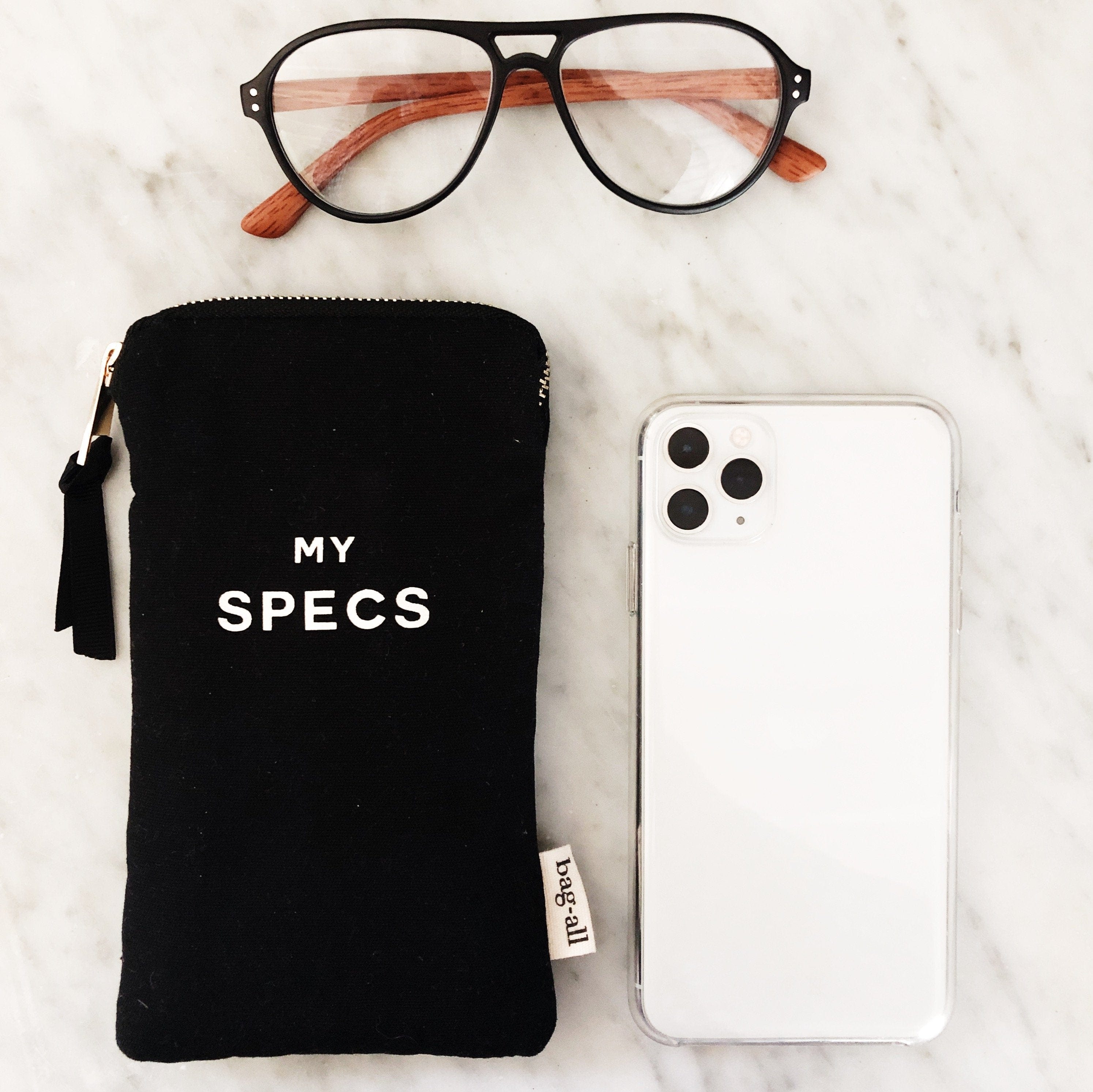 Bag-all My Specs Glasses Case in black canvas with white text, shown with aviator glasses and smartphone, featuring exterior pocket and tassel zipper pull on marble surface