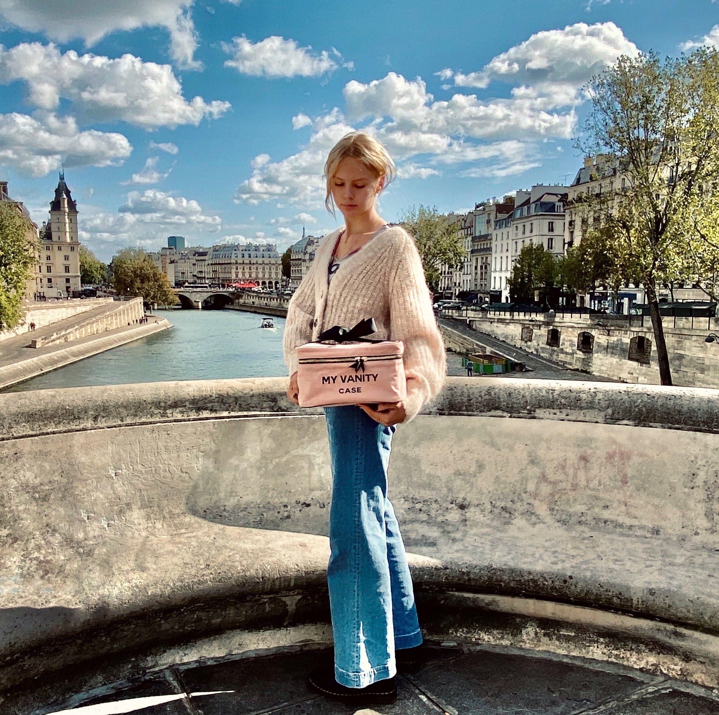 Bag-all My Vanity Large Beauty Box in pink/blush showcased on a scenic Parisian bridge over the Seine River, featuring classic architecture and fluffy white clouds in a bright blue sky