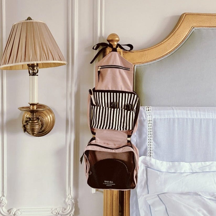 Bag-all Folding/Hanging Toiletry Case in blush pink with striped compartments, featuring multiple storage sections and hanging hook, displayed next to elegant wall sconce lamp and bed headboard