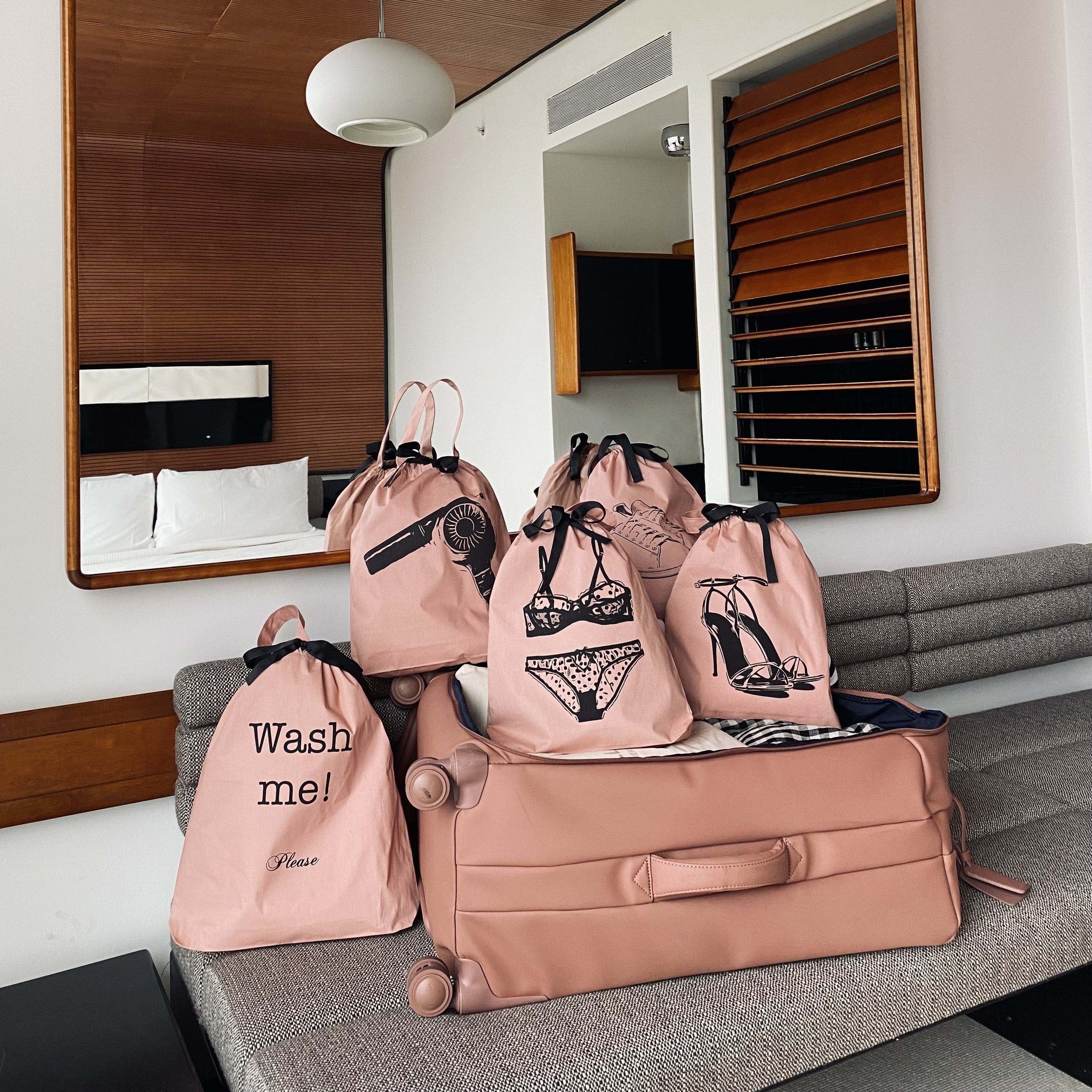 Bag-all pink high heel sandal shoe bag with matching travel organizer bags featuring fashion illustrations and wash me text, displayed on grey couch against mirrored wall