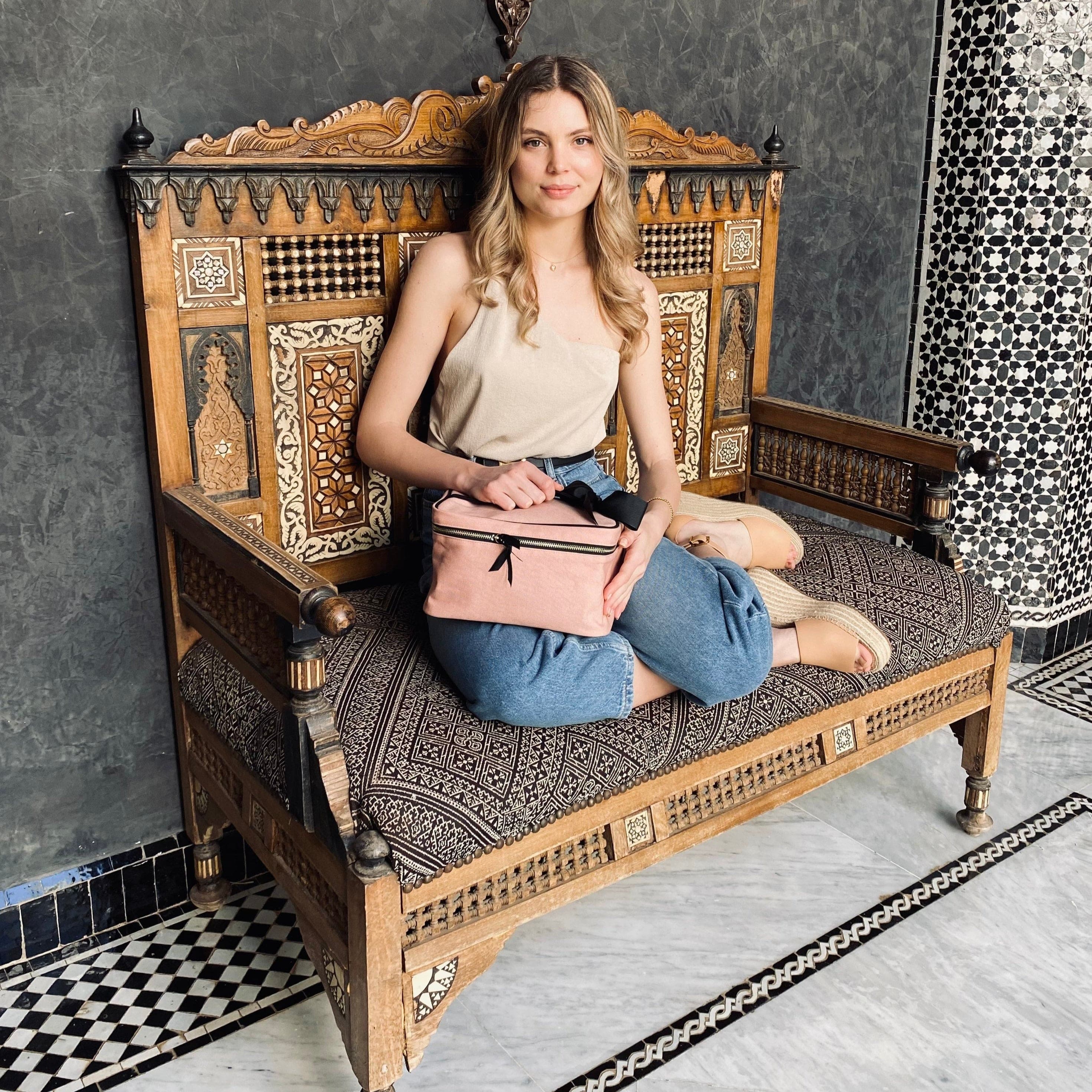 Bag-all Medium Box Makeup & Toiletry in blush pink with black striped lining, stylish bow detail and metal zipper, shown on ornate wooden bench with intricate patterns