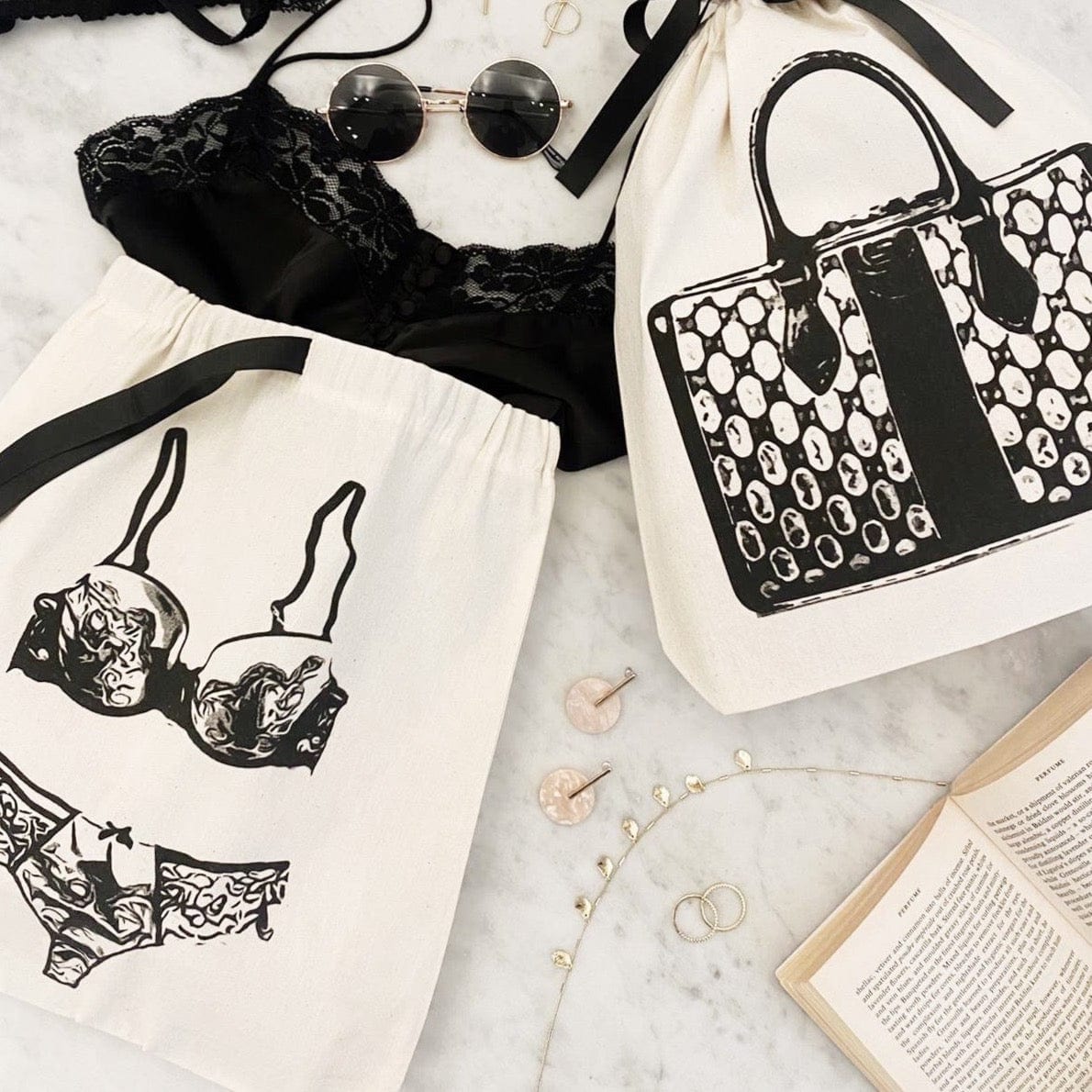 Bag-all Lace Lingerie Travel Bag in cream color with black lingerie illustration, featuring organized storage for delicate garments, displayed with sunglasses and decorative accessories on marble surface