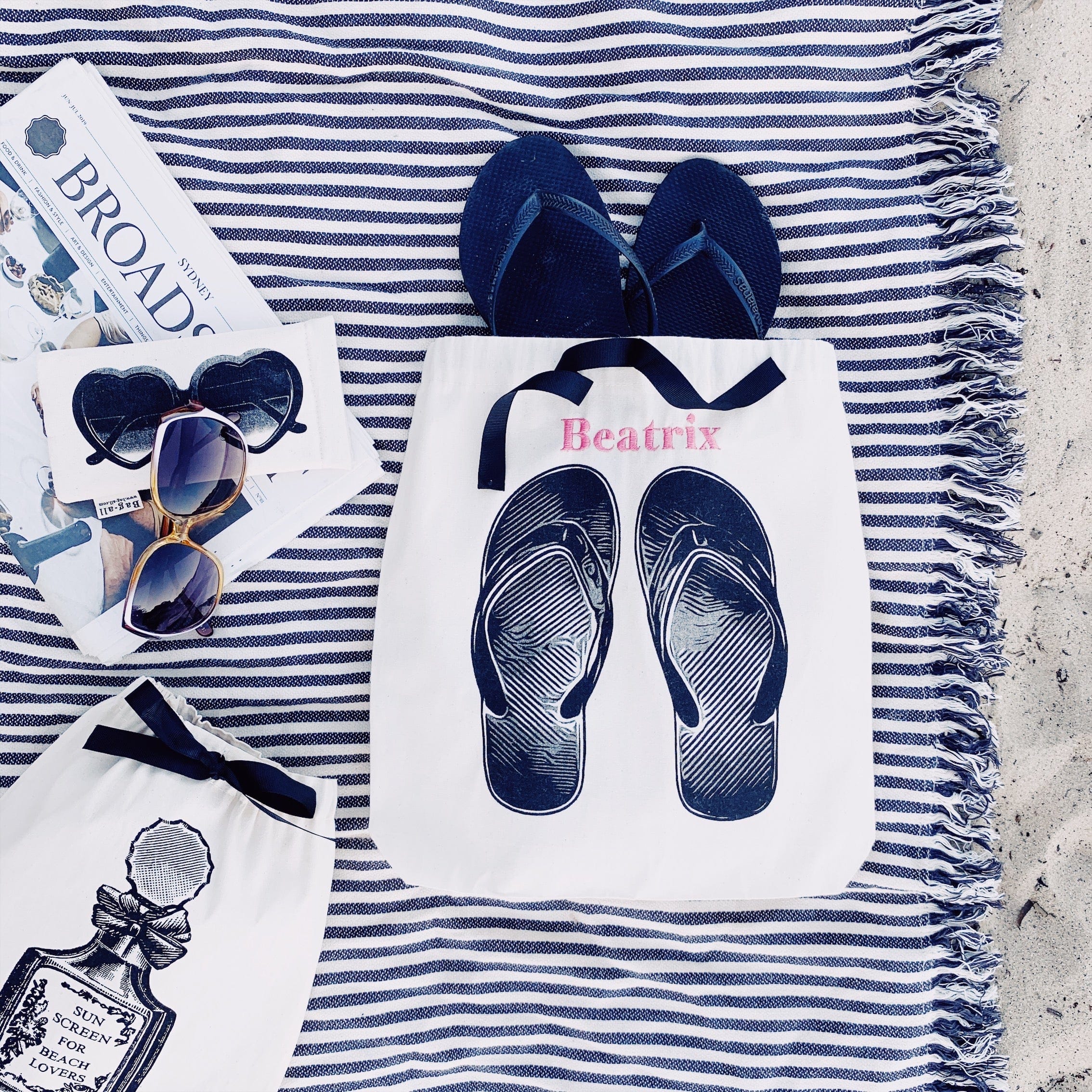 Bag-all Cream Flip Flops Shoe Bag with navy ribbon accent shown on striped beach blanket with sunglasses and travel accessories, perfect for beach organization and summer storage
