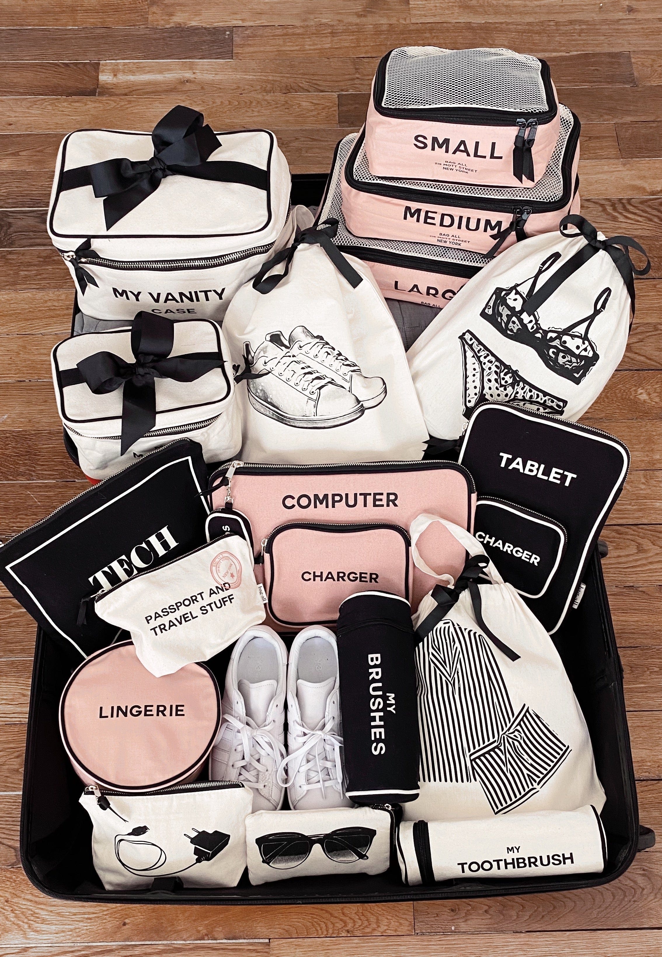 Bag-all black toothbrush travel case displayed with collection of branded organizing bags in pink, black and white featuring labeled compartments for tech, toiletries and clothing essentials on wooden floor