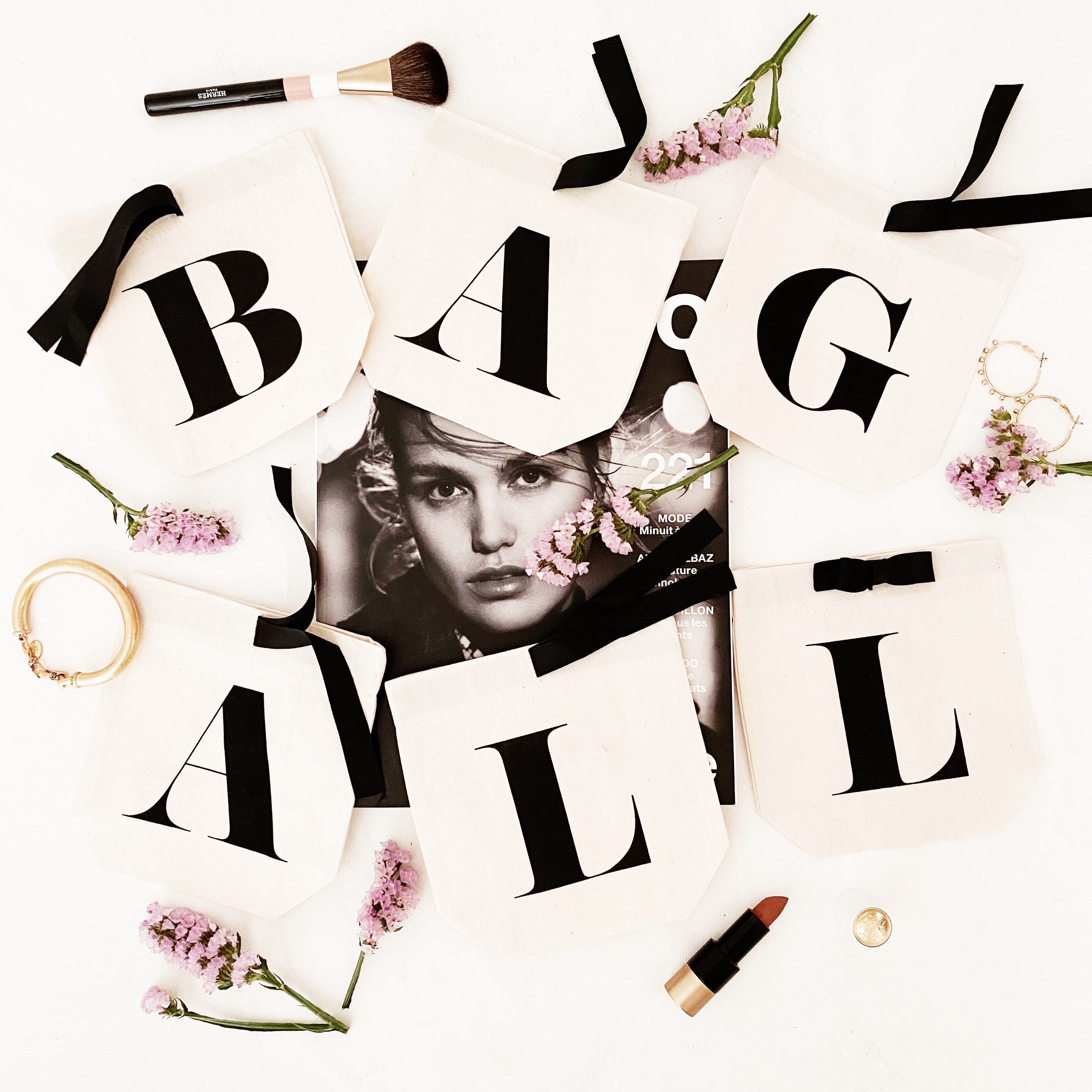 Bag-all Small Letter Bags Classic in cream with black typography letters, arranged with pink flowers, makeup brushes and lipstick on white background, perfect for organizing essentials