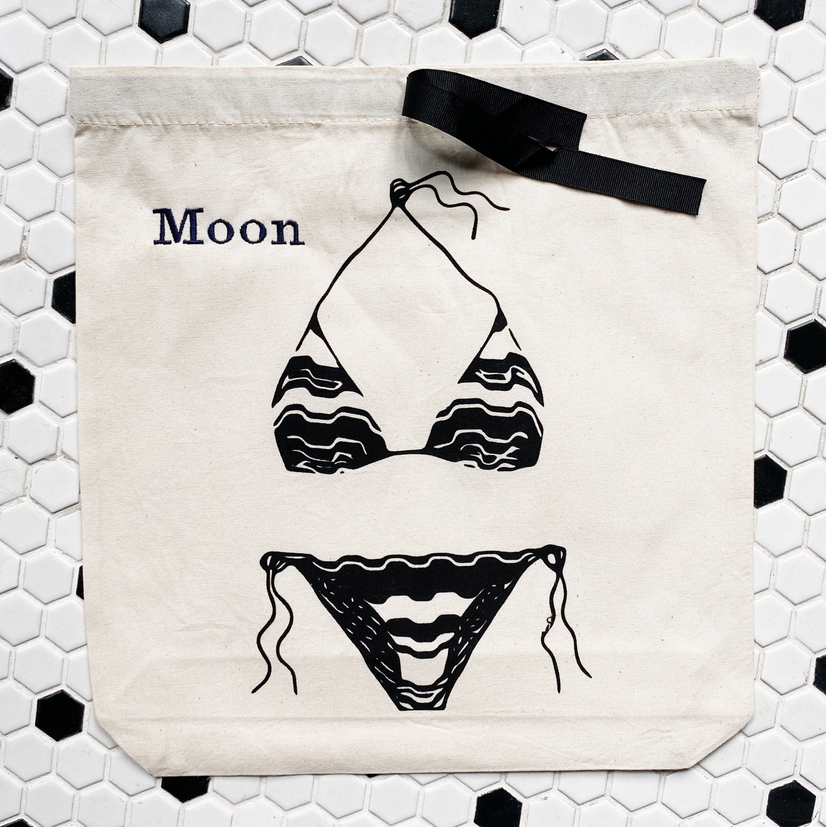 Bag-all Triangle Bikini Travel Bag in cream cotton featuring black bikini illustration and Moon text, with drawstring closure against hexagonal tile background