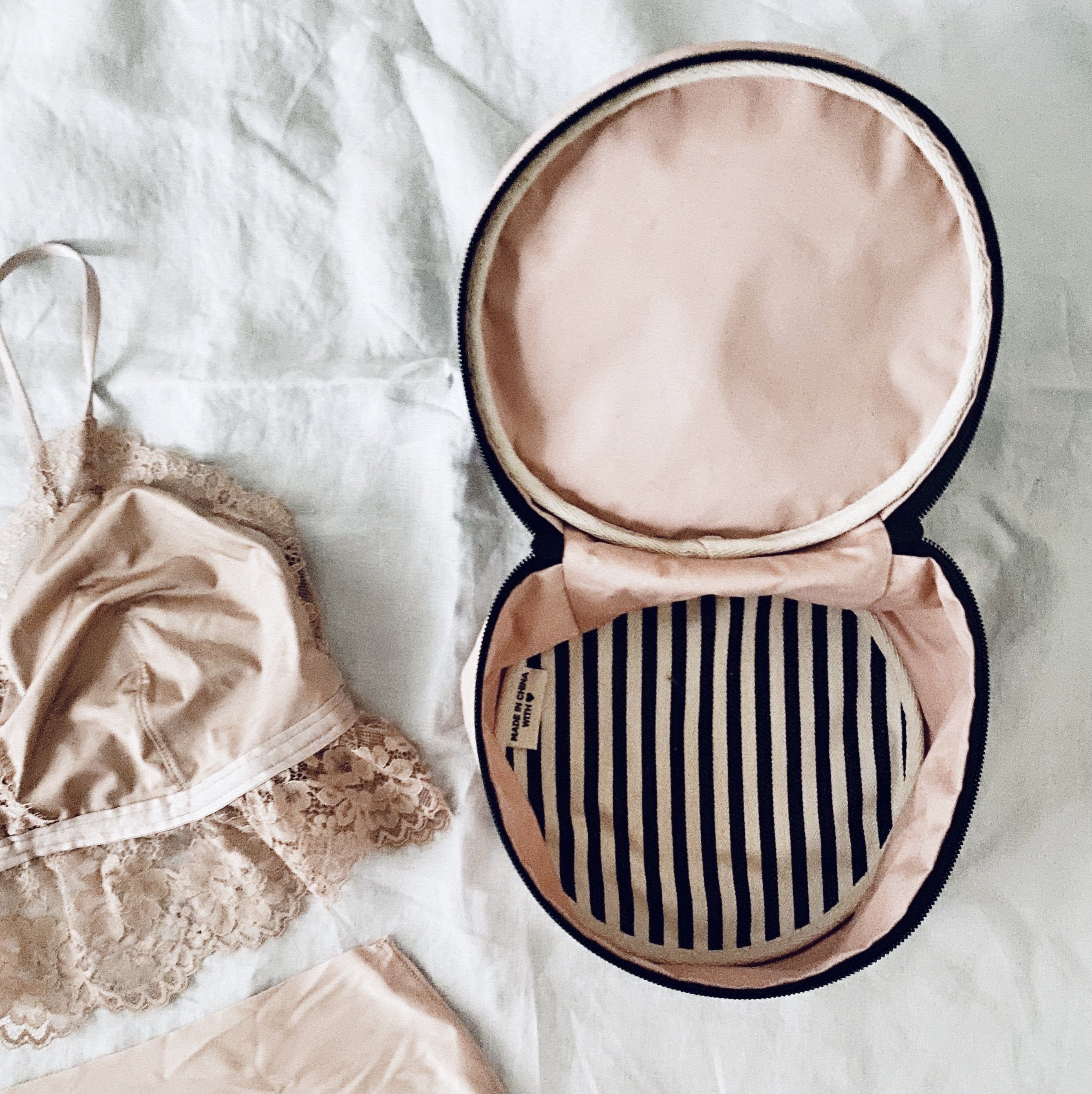 Bag-all Round Lingerie Case in blush pink with black and white striped interior lining, shown open next to delicate lace undergarments on white background
