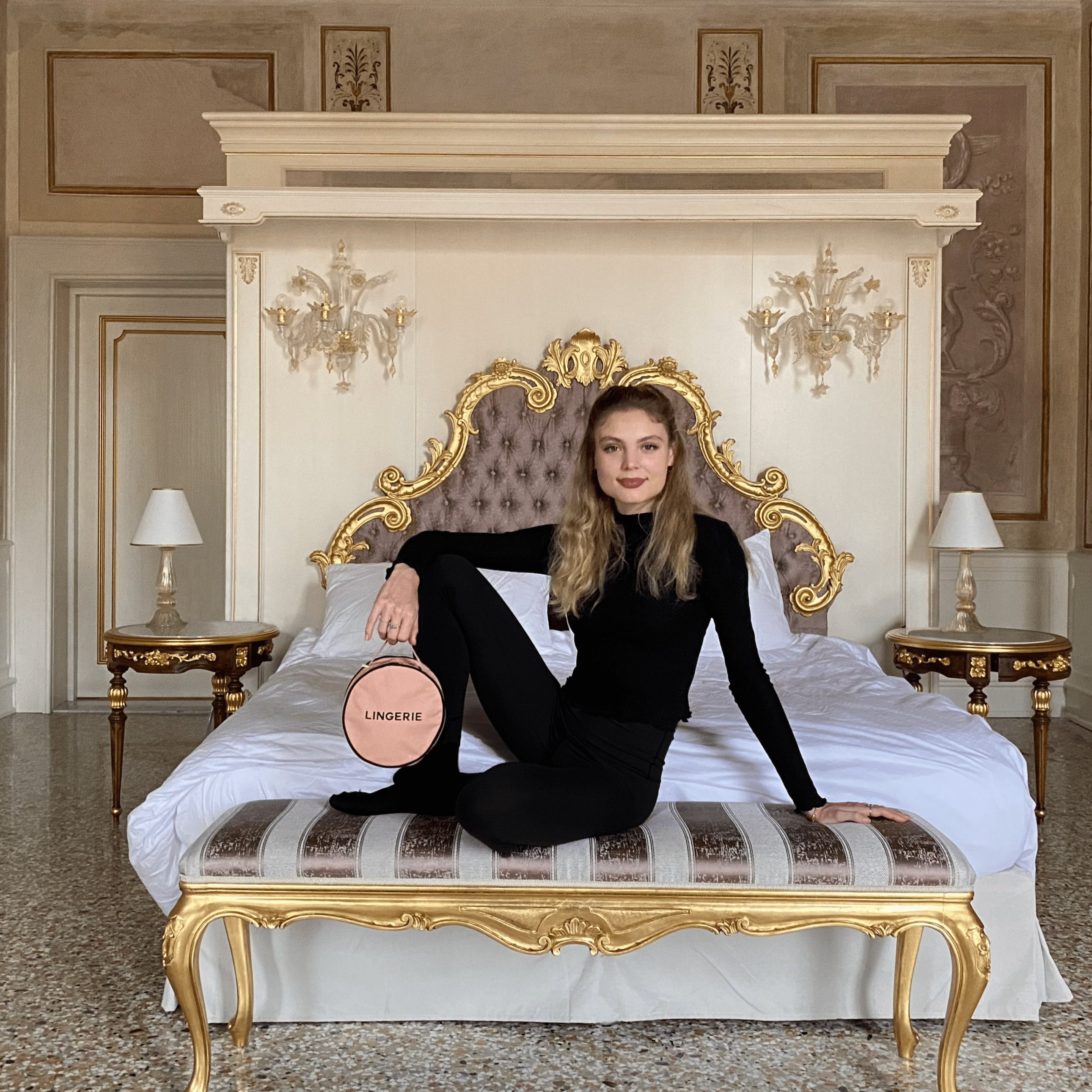 Bag-all Round Lingerie Case in blush pink shown on ornate vintage bed with elegant baroque headboard and crystal wall sconces in luxurious bedroom setting
