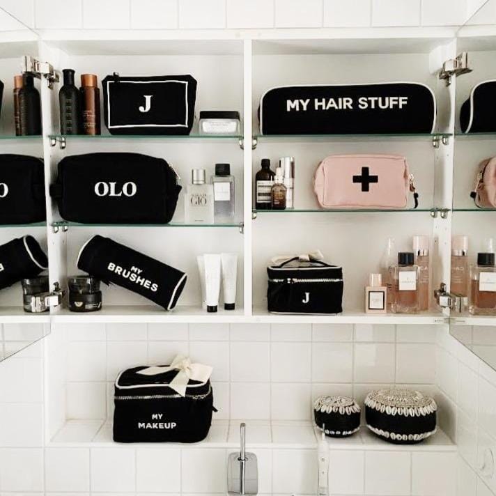 White bathroom shelving styled with black fabric travel organizers and cosmetic cases labeled “My Makeup,” “My Hair Stuff,” and “My Brushes,” arranged alongside skincare bottles, perfumes, and toiletry items for an organized vanity display.