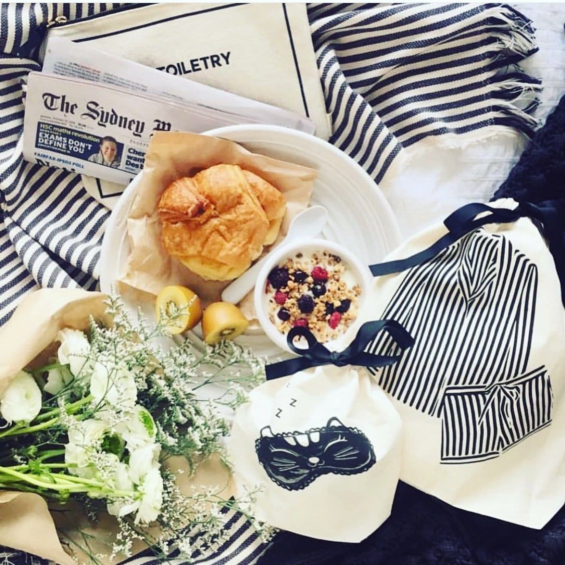 Bag-all Striped Pajamas Travel Bag in cream, styled with breakfast, fresh flowers, and newspaper, showcasing classic navy and white stripes against white bedding, perfect for travel organization