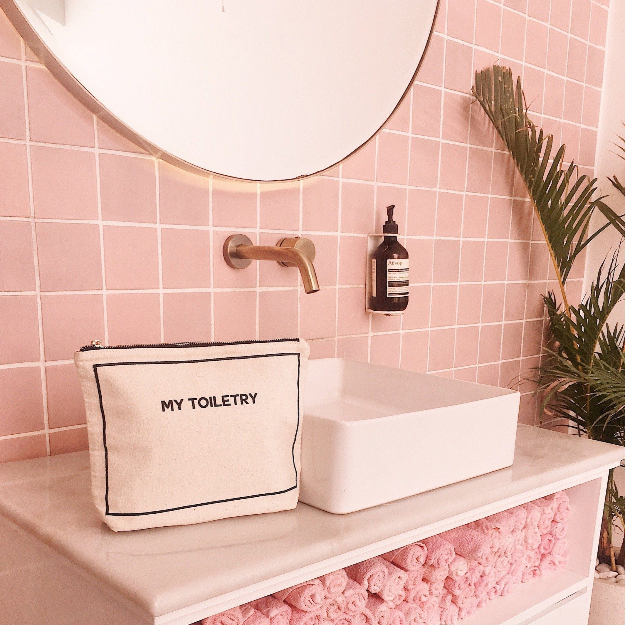 Bag-all Toiletry Pouch with Coated Lining in cream canvas sits elegantly on modern bathroom counter with pink tile backdrop, featuring black embroidered text and stylish leak-proof design