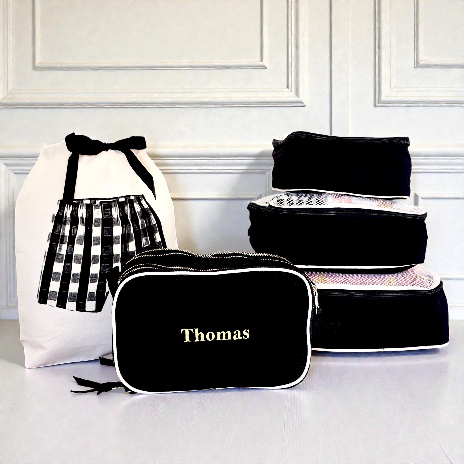 Black personalized travel organizer and 5-pack packing cubes by Bag-all stacked beside white laundry bag with bow detail