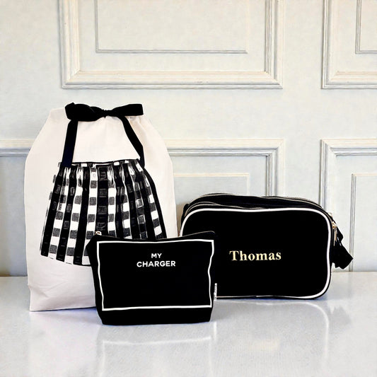 Black personalized toiletry, tech, and travel organizers set by Bag-all with white trim and gold zipper accents on white surface