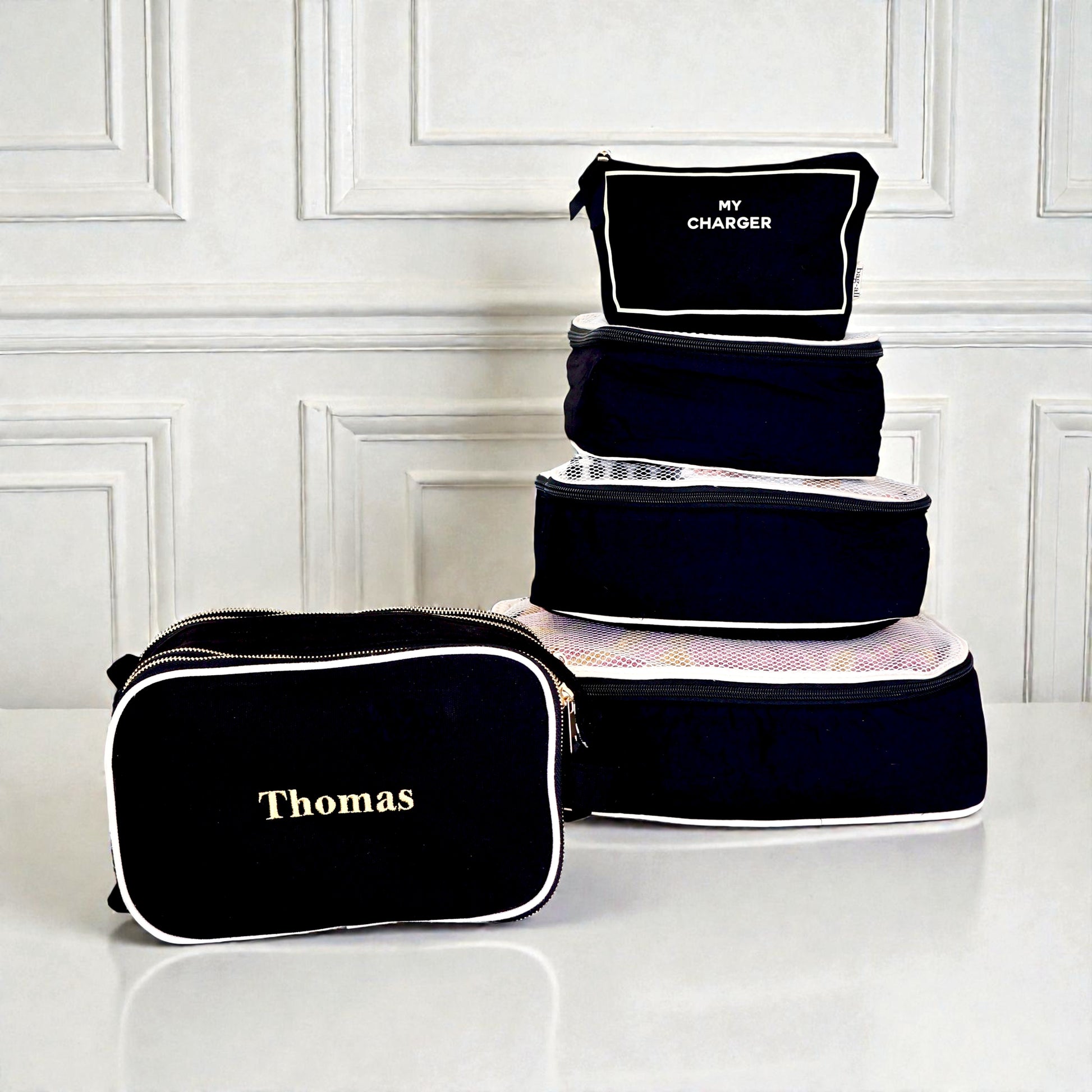 Five black Bag-all packing cubes and organizers with white piping and personalized text on smooth surface