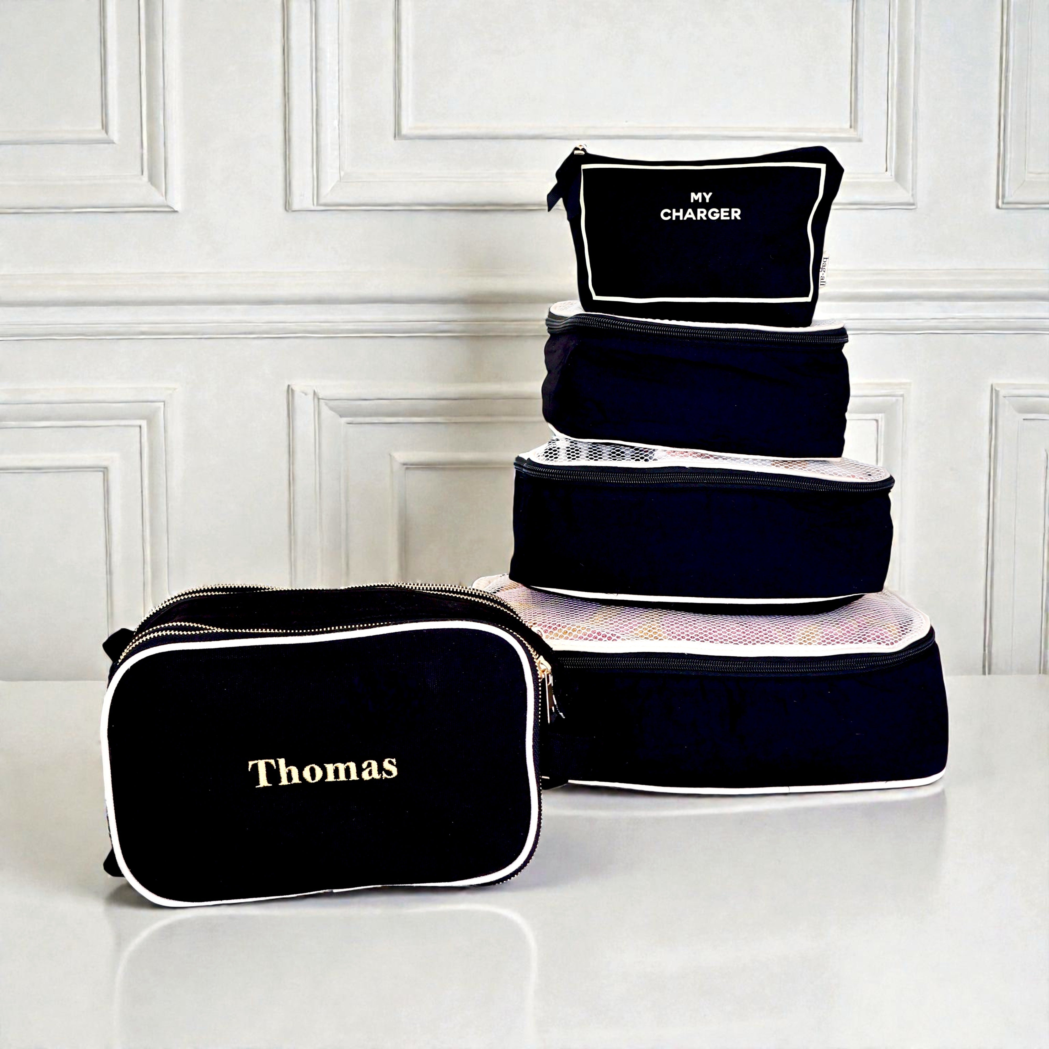 Five black Bag-all packing cubes and organizers with white piping and personalized text on smooth surface