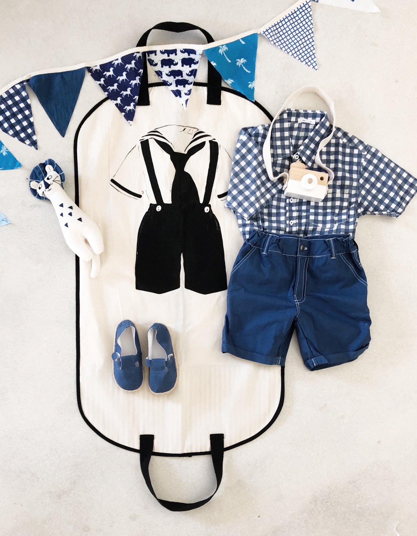Bag-all Kids Garment Bag Boy in cream featuring stylish black suspender design, with convenient handles and pockets, displayed with blue gingham outfit and decorative bunting