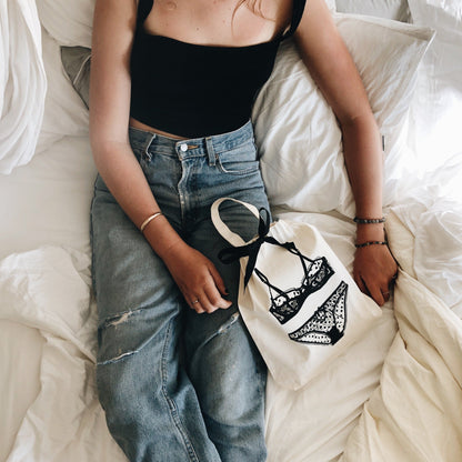 Woman in black top and ripped jeans holding cream lingerie organizer with black ribbon by Bag-all on white bedding