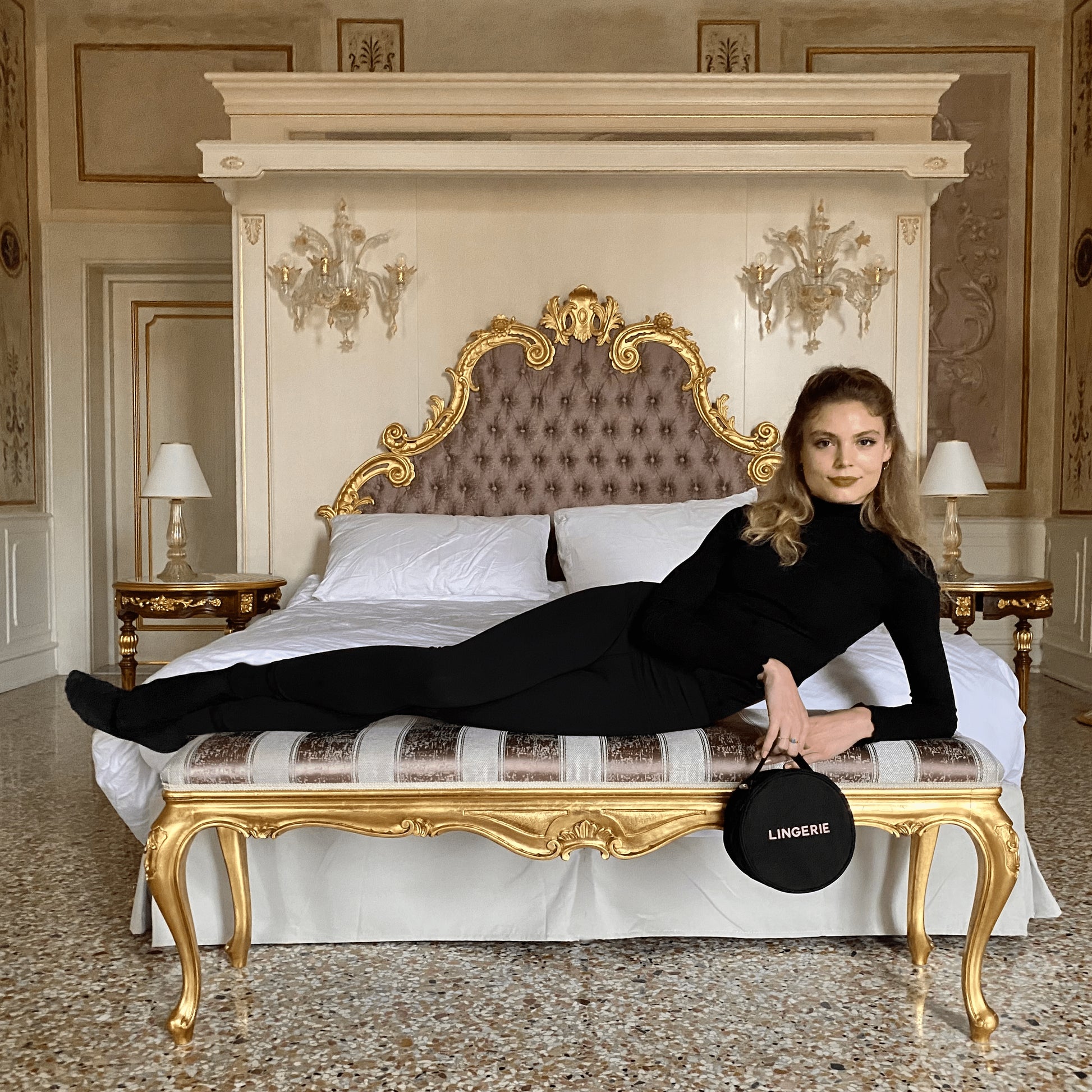Woman dressed in black reclining on ornate bench holding black round lingerie case with Bag-all branding on bed background