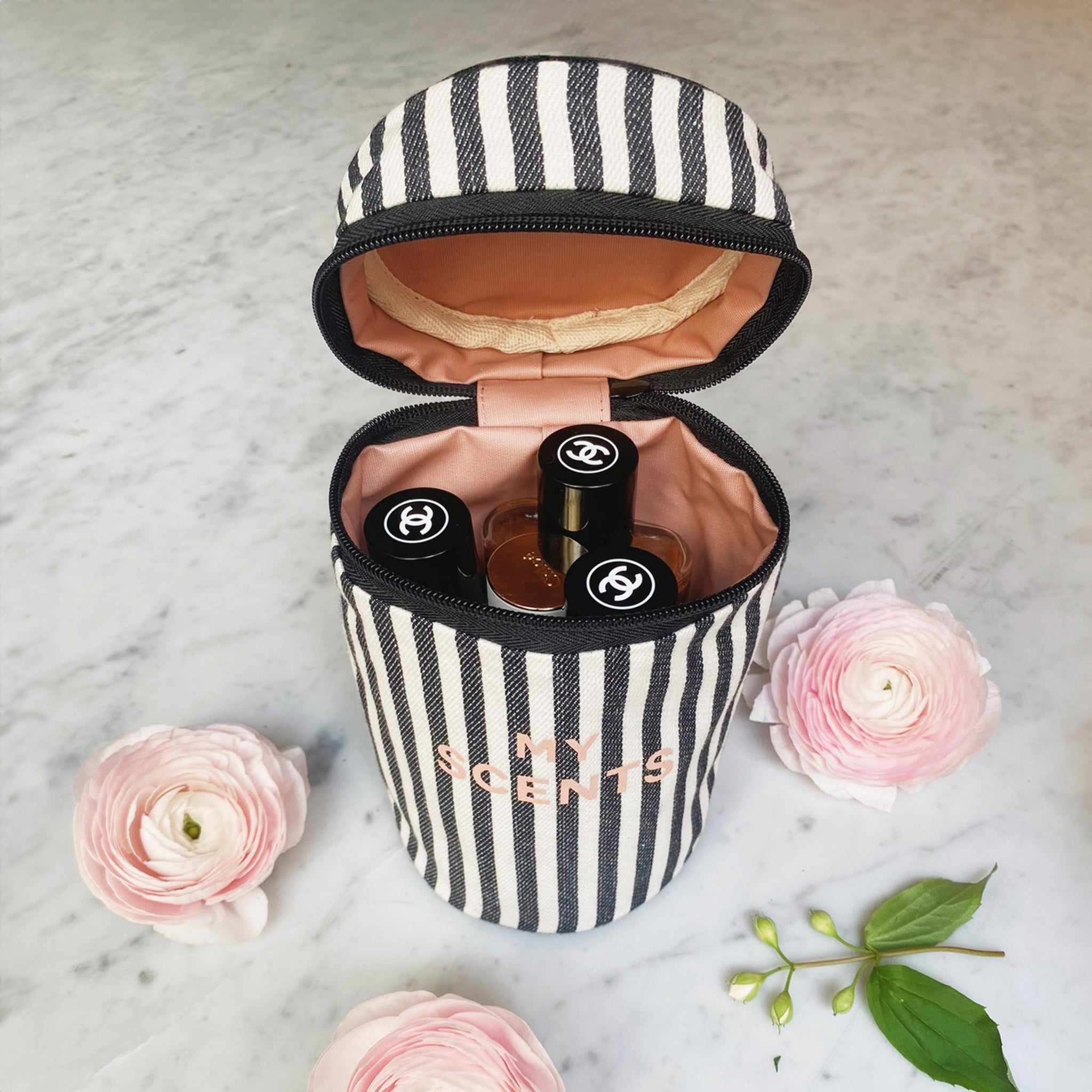 Bag-all Round My Scents Case in black and white stripes, opened to display perfume storage compartment with pink interior lining, surrounded by delicate pink ranunculus flowers on marble surface
