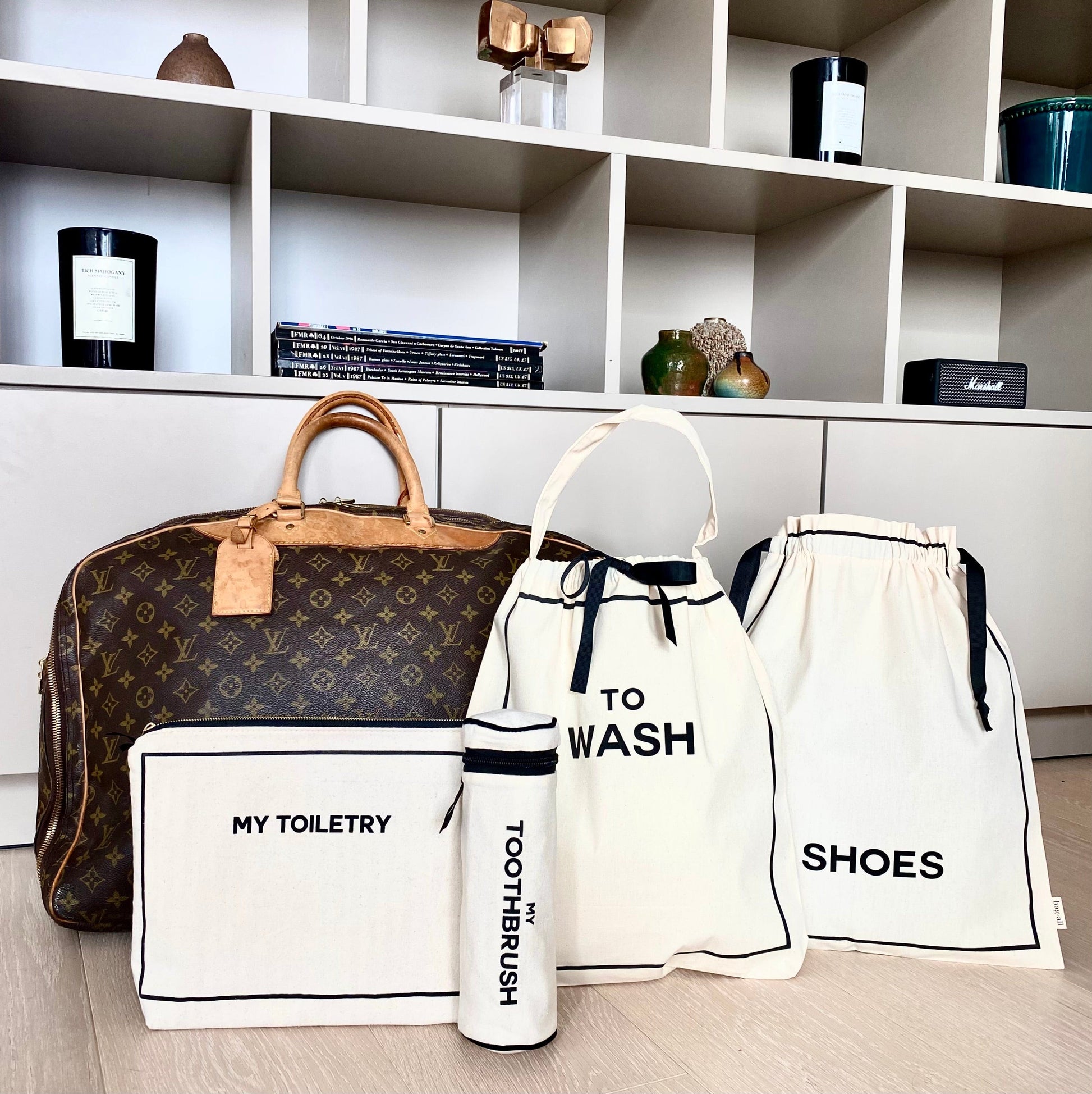 Bag-all Toiletry Travel Gift Set 4-Pack in cream featuring cotton organizing pouches for toiletries, toothbrush, shoes, and washing items displayed next to designer travel bag on white shelf unit