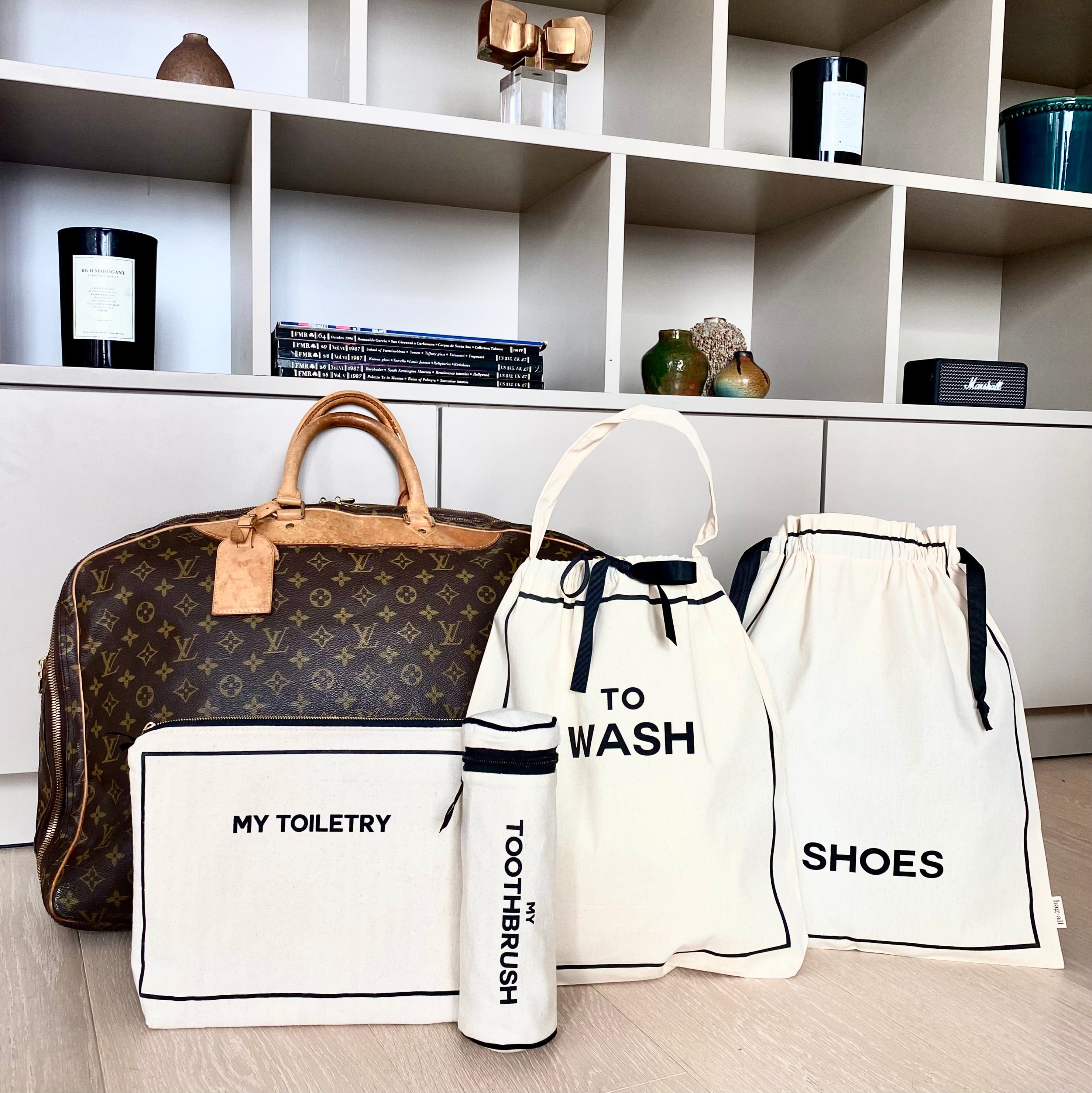 Bag-all Toiletry Travel Gift Set 4-Pack in cream featuring cotton organizing pouches for toiletries, toothbrush, shoes, and washing items displayed next to designer travel bag on white shelf unit