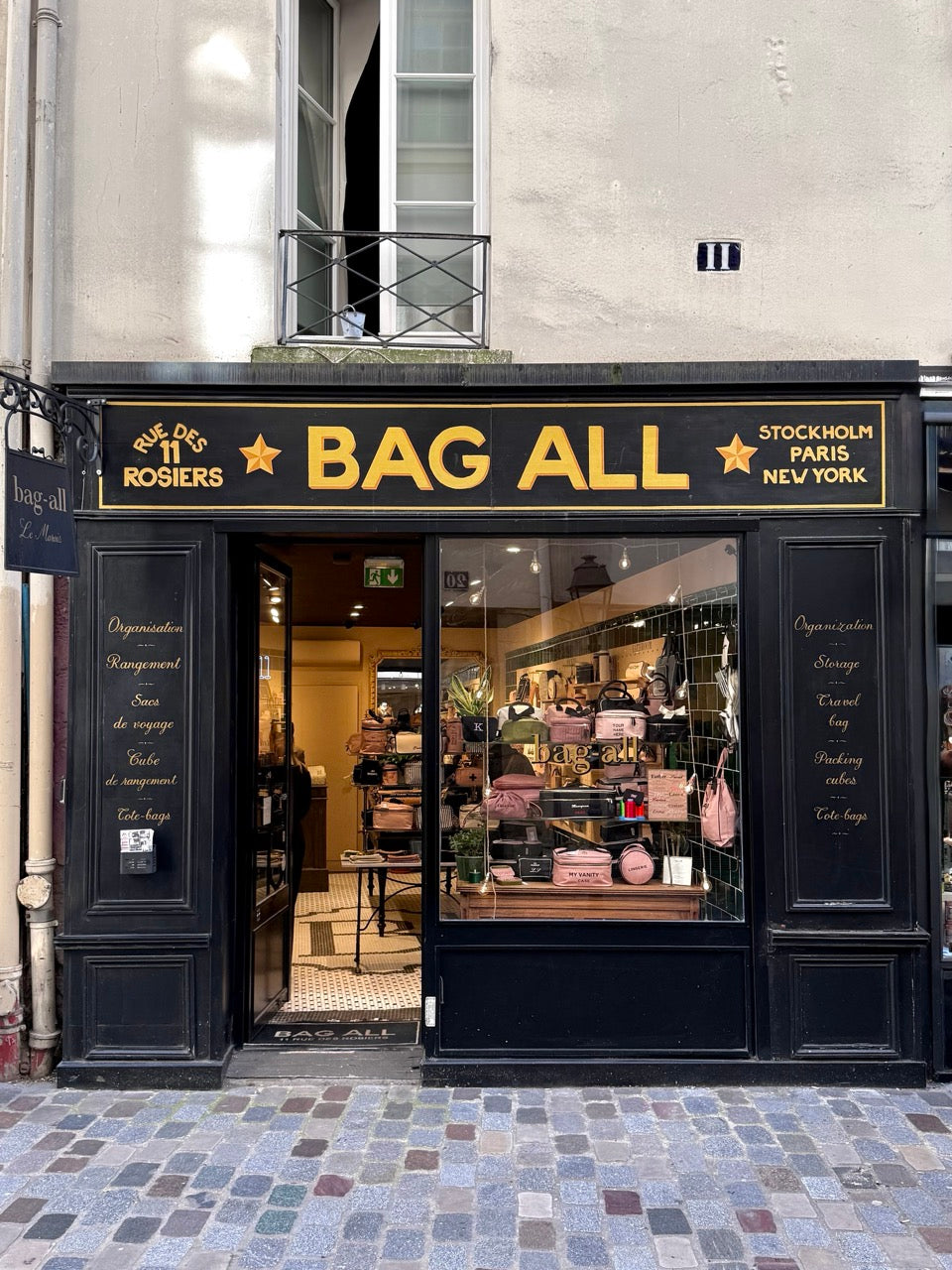 Bag-all boutique storefront with black and gold sign, displaying organized pink bags inside on cobblestone street gallery position 1