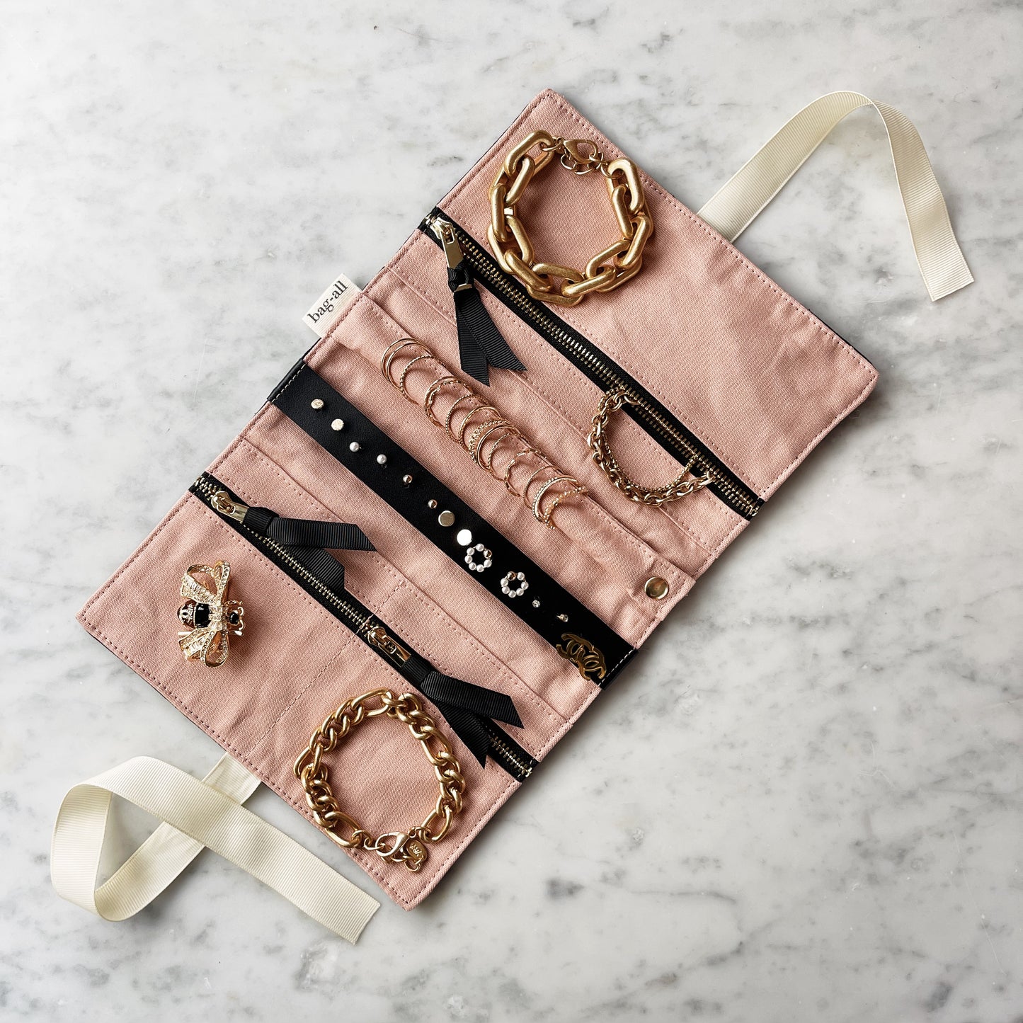 Bag-all Couture Jewelry Roll Medium in black featuring pink interior, multiple compartments, ribbon ties, and chain jewelry displayed elegantly on marble surface