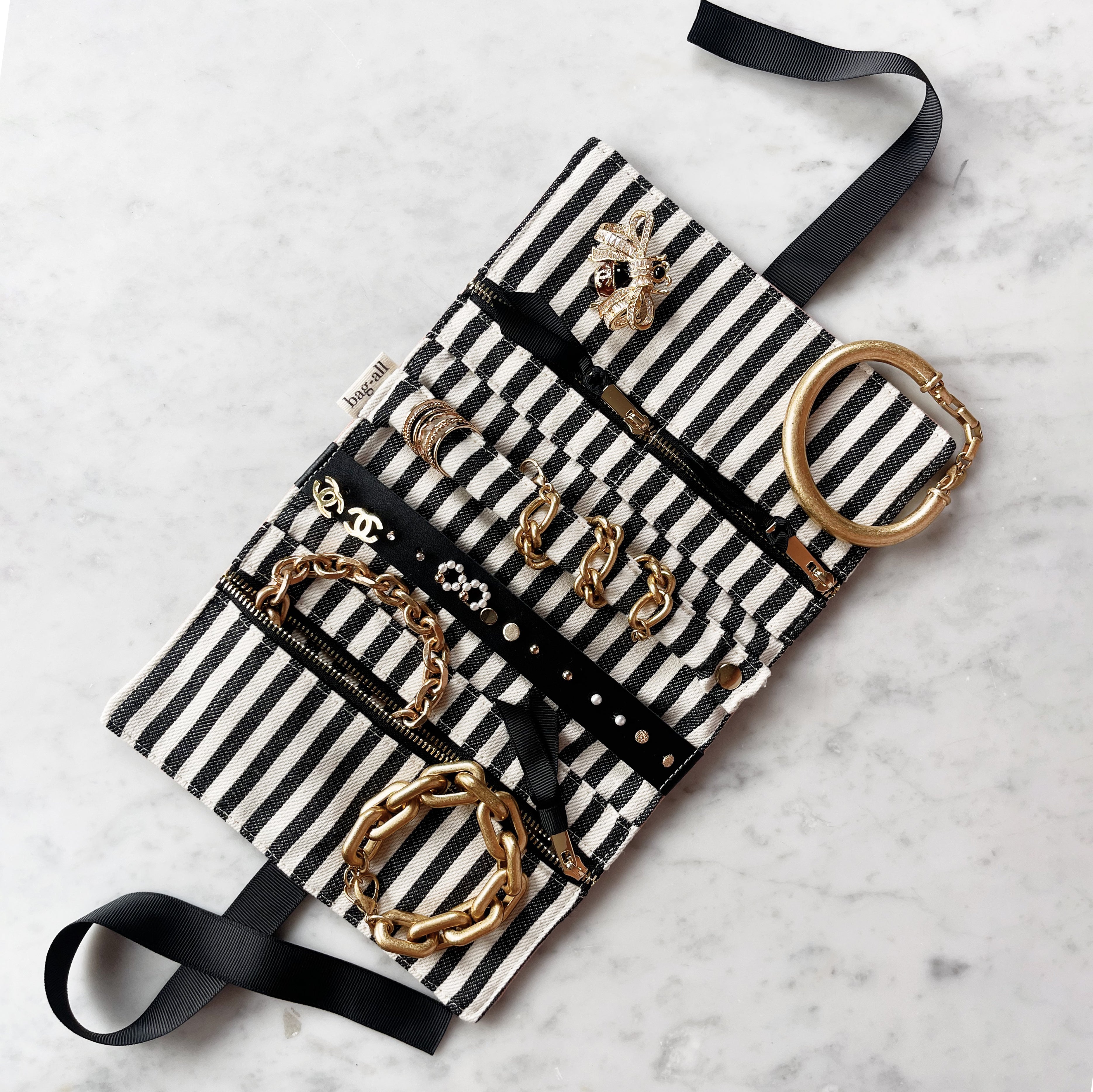 Bag-all Couture Jewelry Roll Medium in pink with black and white striped interior, featuring multiple compartments displaying organized gold jewelry and accessories on marble surface
