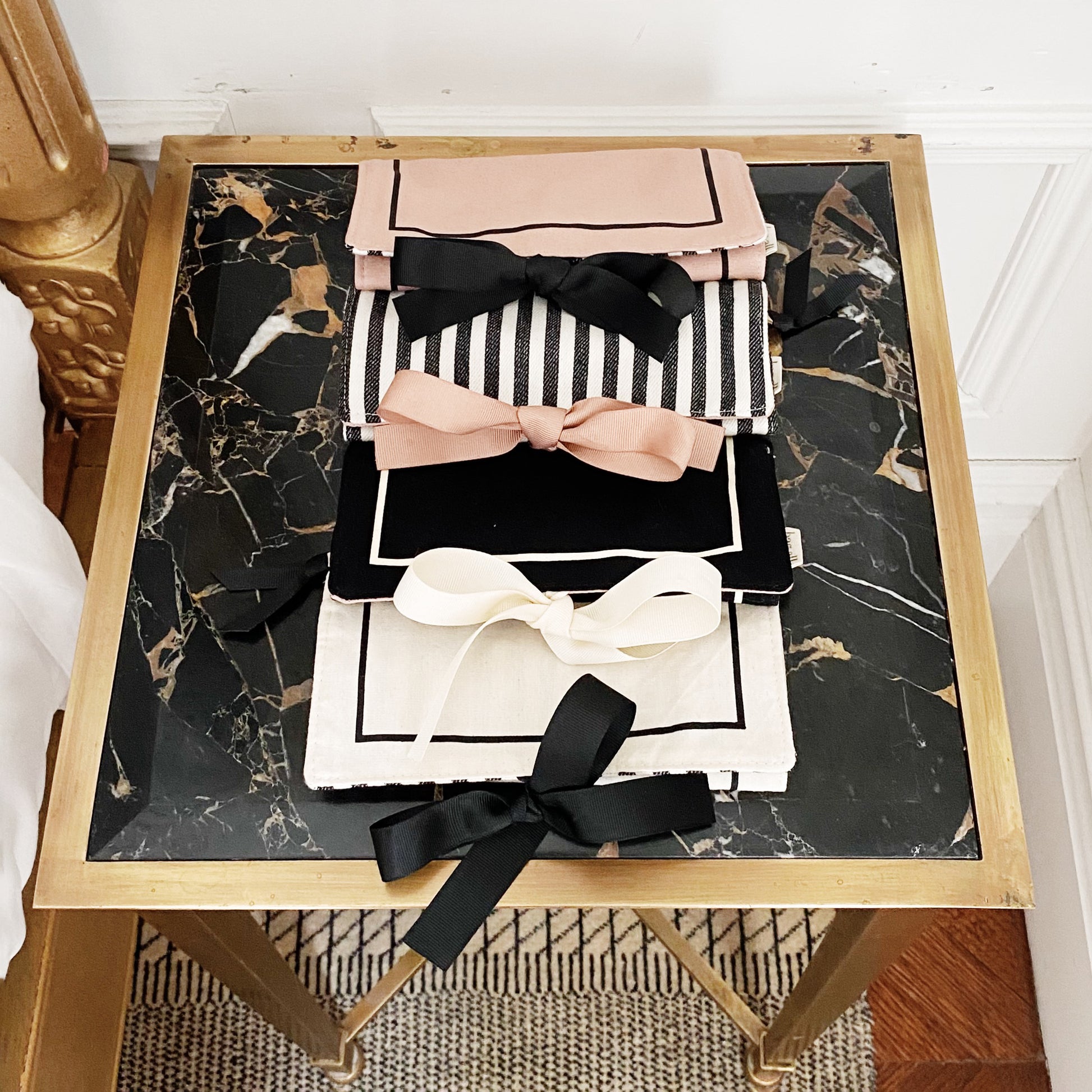 Bag-all Couture Jewelry Roll Medium Black: Elegant travel organizer with decorative bows in pink, black, and white, displayed on marble-top gold table, featuring striped pattern and multiple compartments