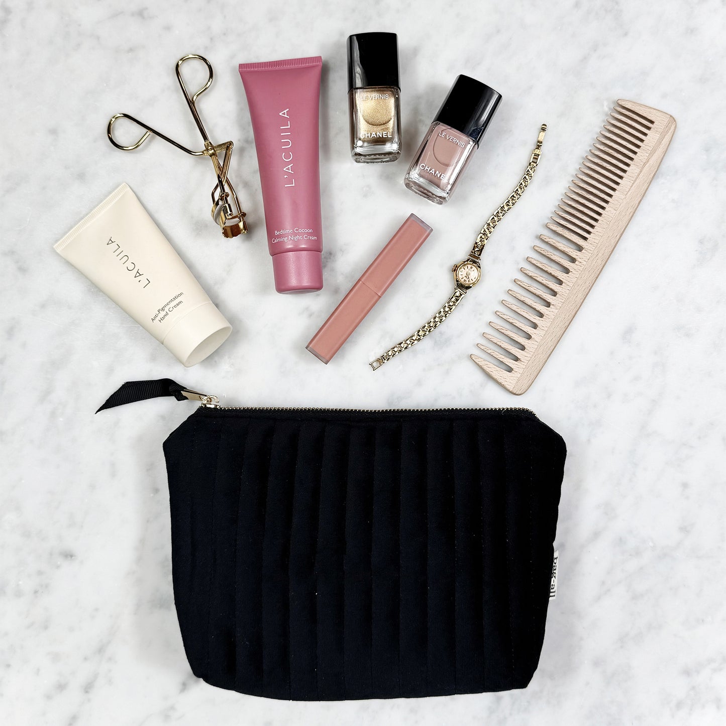 Black quilted velvet Bag-all pouch with makeup, nail polish, watch, comb, and eyelash curler on marble surface