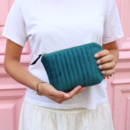 Woman holding quilted teal velvet makeup pouch with gold zipper and Bag-all branding in pink background
