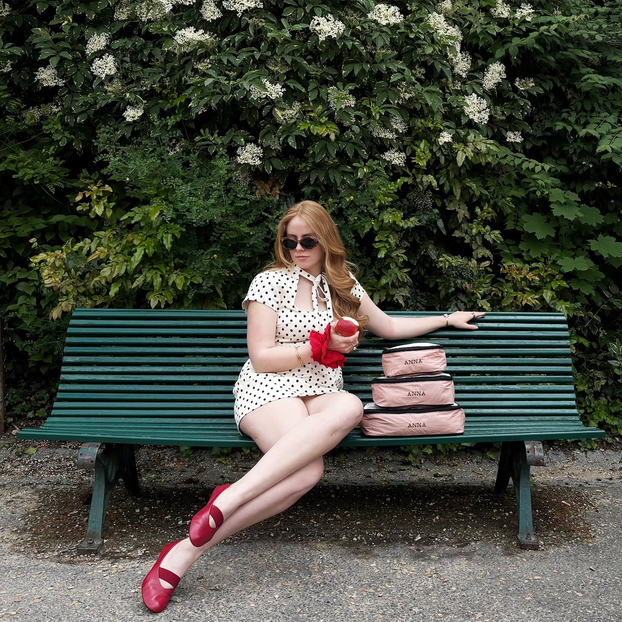 Woman in polka dot dress wearing sunglasses seated on green bench with pink personalized Bag-all cotton travel organizers for stylish packing.