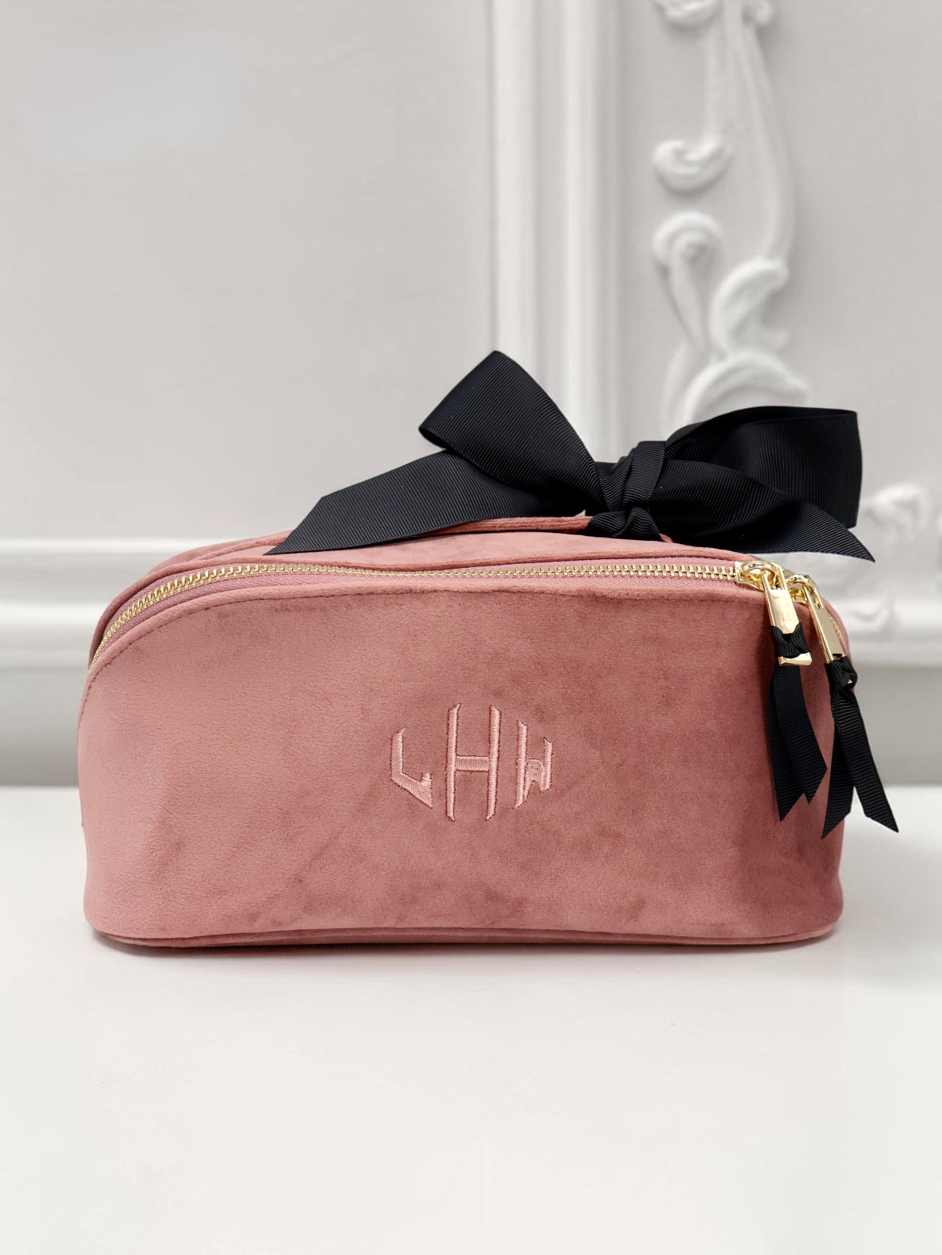 Blush pink velvet travel pouch with embroidered monogram and gold zipper accented by black ribbon bows, reflecting Bag-all’s Paris-designed style.