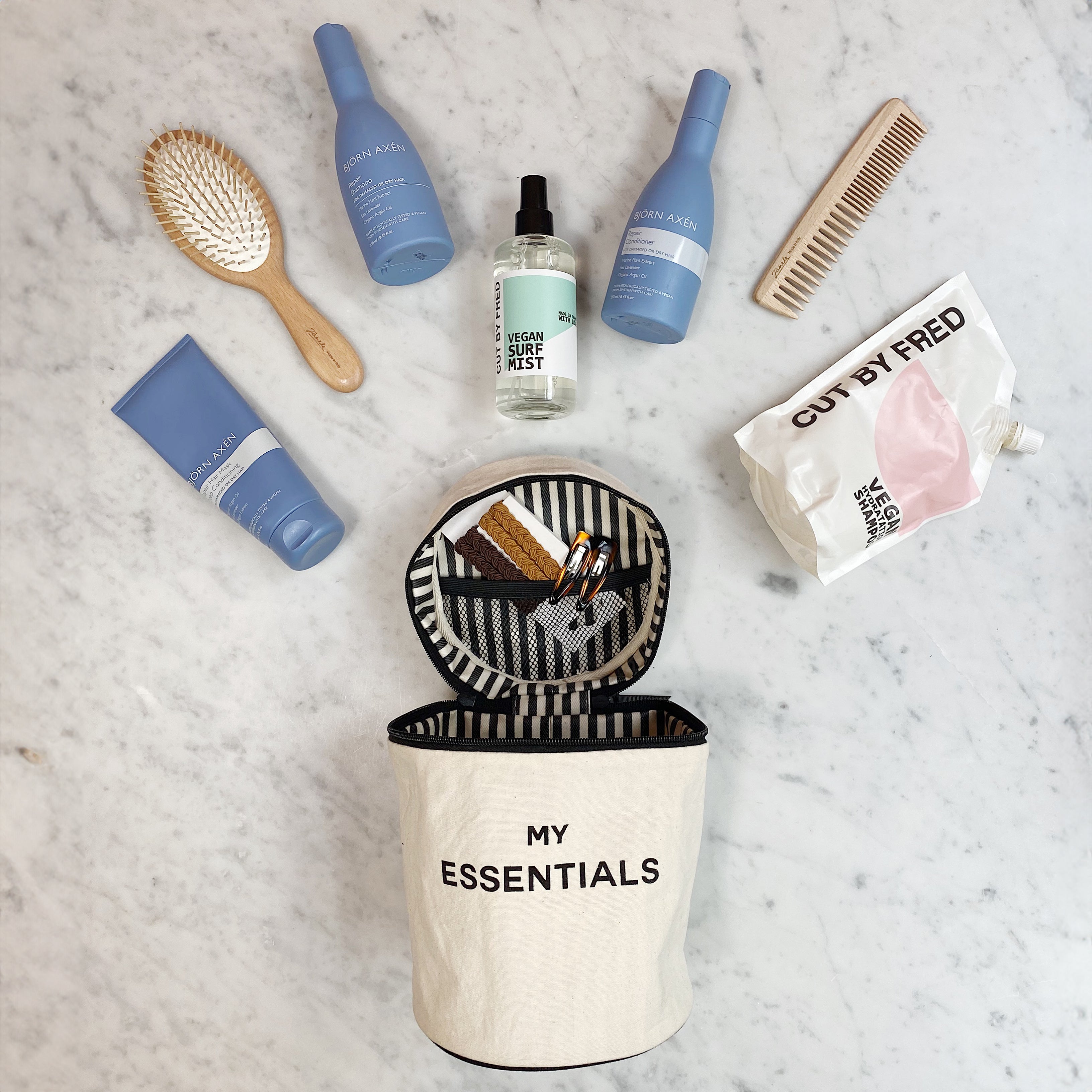 Bag-all Roomy Travel Essentials Case in cream with striped lid, shown open displaying various beauty products including blue bottles, hairbrush, and spray bottle on marble surface