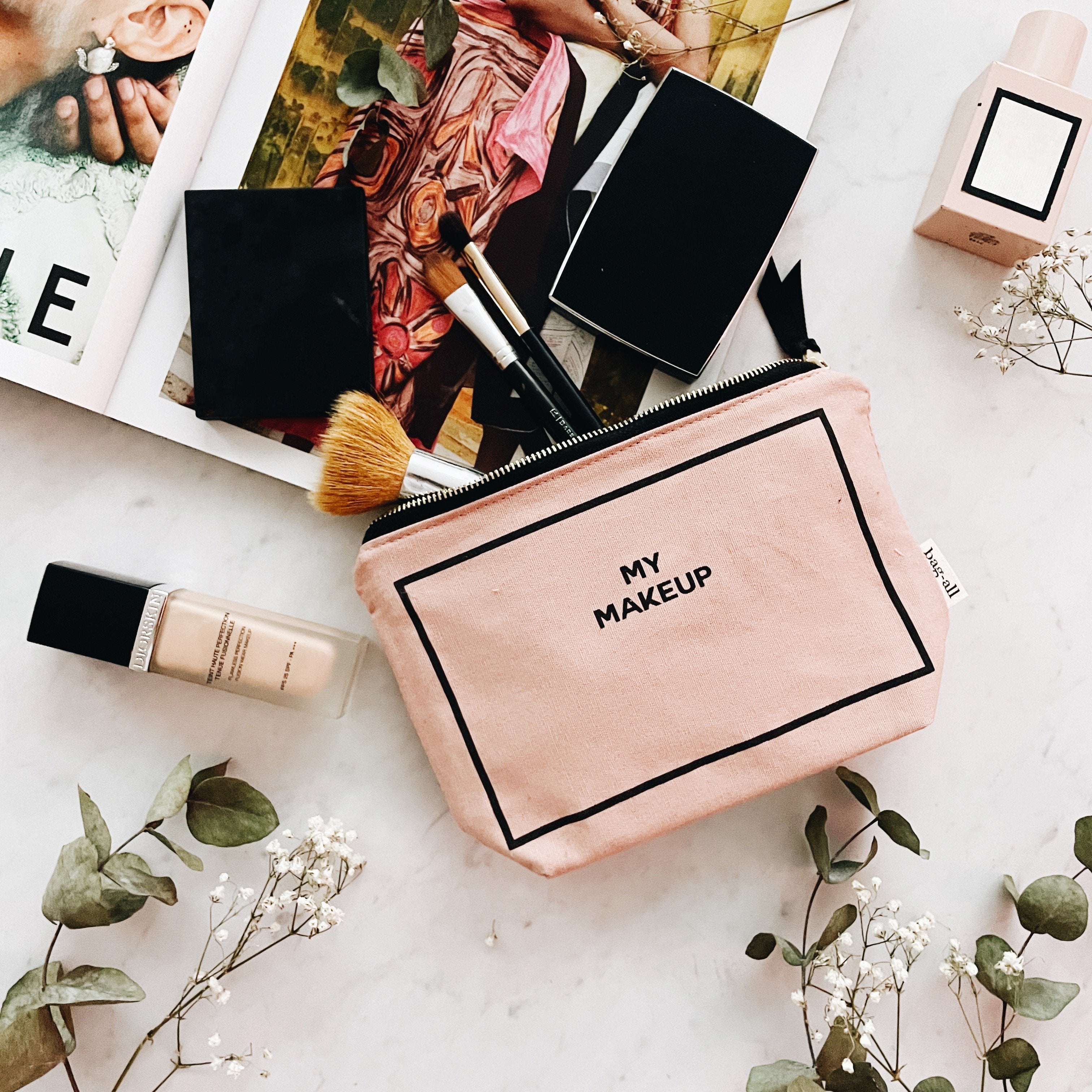 Bag-all pink blush makeup pouch with black trim and zipper holding brushes on marble surface with floral and beauty items