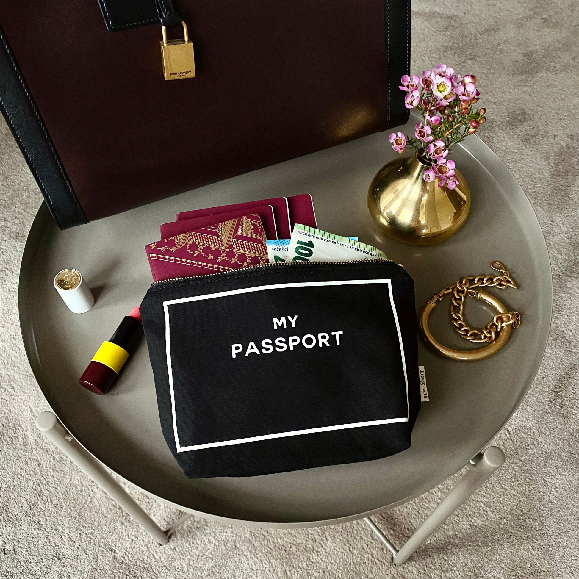 Bag-all black passport organizer pouch on table with travel essentials including money and lipstick in modern setting