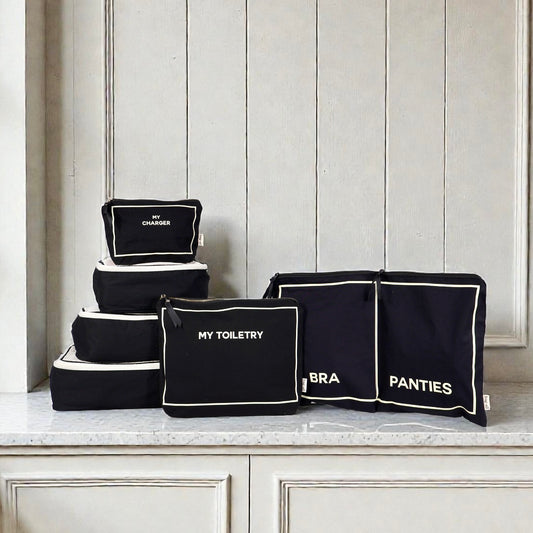 Set of black Bag-all travel packing cubes labeled My Charger, My Toiletry, Bra, and Panties on marble surface